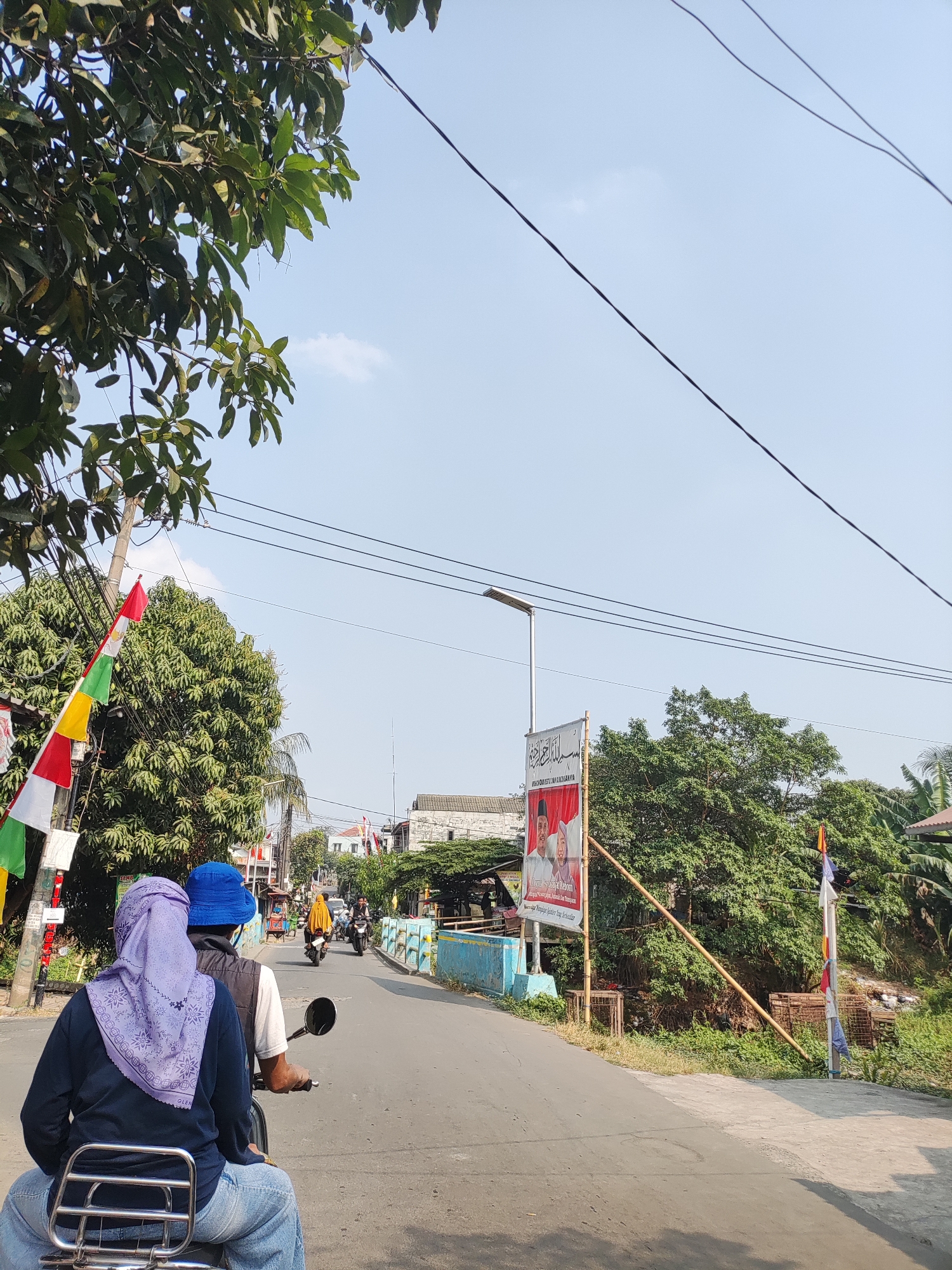 A street scene in an outdoor, possibly rural, environment is depicted under a clear, bright sky. The perspective is from behind a person riding on the back of a scooter or motorcycle. In the foreground, the back of a person wearing a blue helmet and a light purple headscarf is visible, with another person in front. They are on a street that appears to be paved. Several colorful flags, red, white, green, and yellow, are attached to a utility pole on the left side of the road. Trees with dense green foliage line the sides of the street. Further down the road, other motorcycles and a few buildings are visible. A large banner with images of two people and text in Arabic script is displayed on the right side of the street. Overhead power lines are strung across the upper portion of the image. The overall impression is a sunny day in a populated area with elements of everyday life and possibly a local event or election campaign indicated by the flags and banner.
