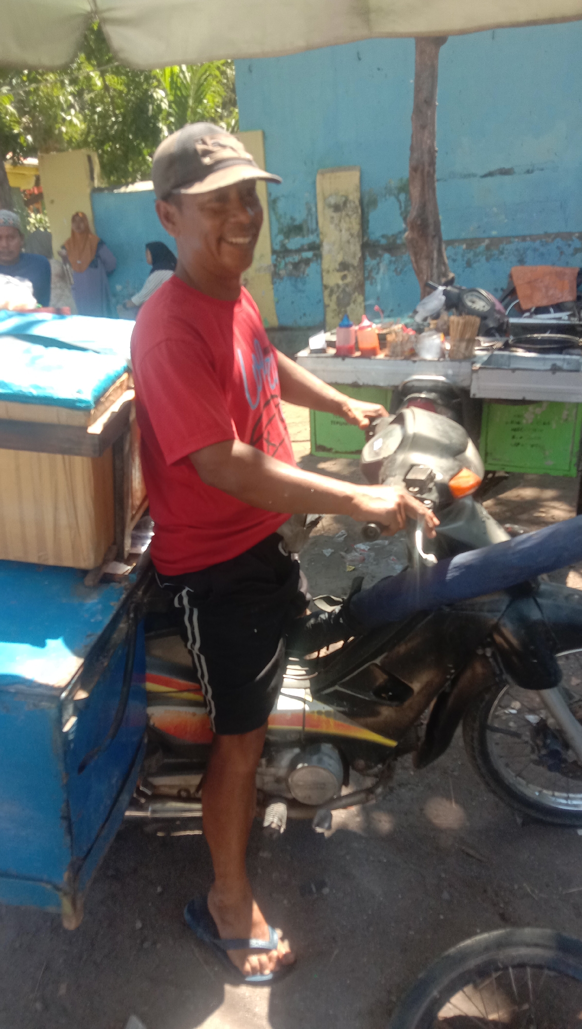 A man wearing a red t-shirt, black shorts with white stripes, a brown baseball cap, and blue flip-flops stands next to a motorcycle with a sidecar attached. The motorcycle is parked on a dirt road in what appears to be a market stall area. In the background, there are other people, a blue wall, and some trees. The sidecar of the motorcycle has a wooden box on top covered with a blue tarp. The man is smiling and looking towards the right side of the frame. The scene is lit by bright sunlight. The location is identified as Ngempit, Indonesia.