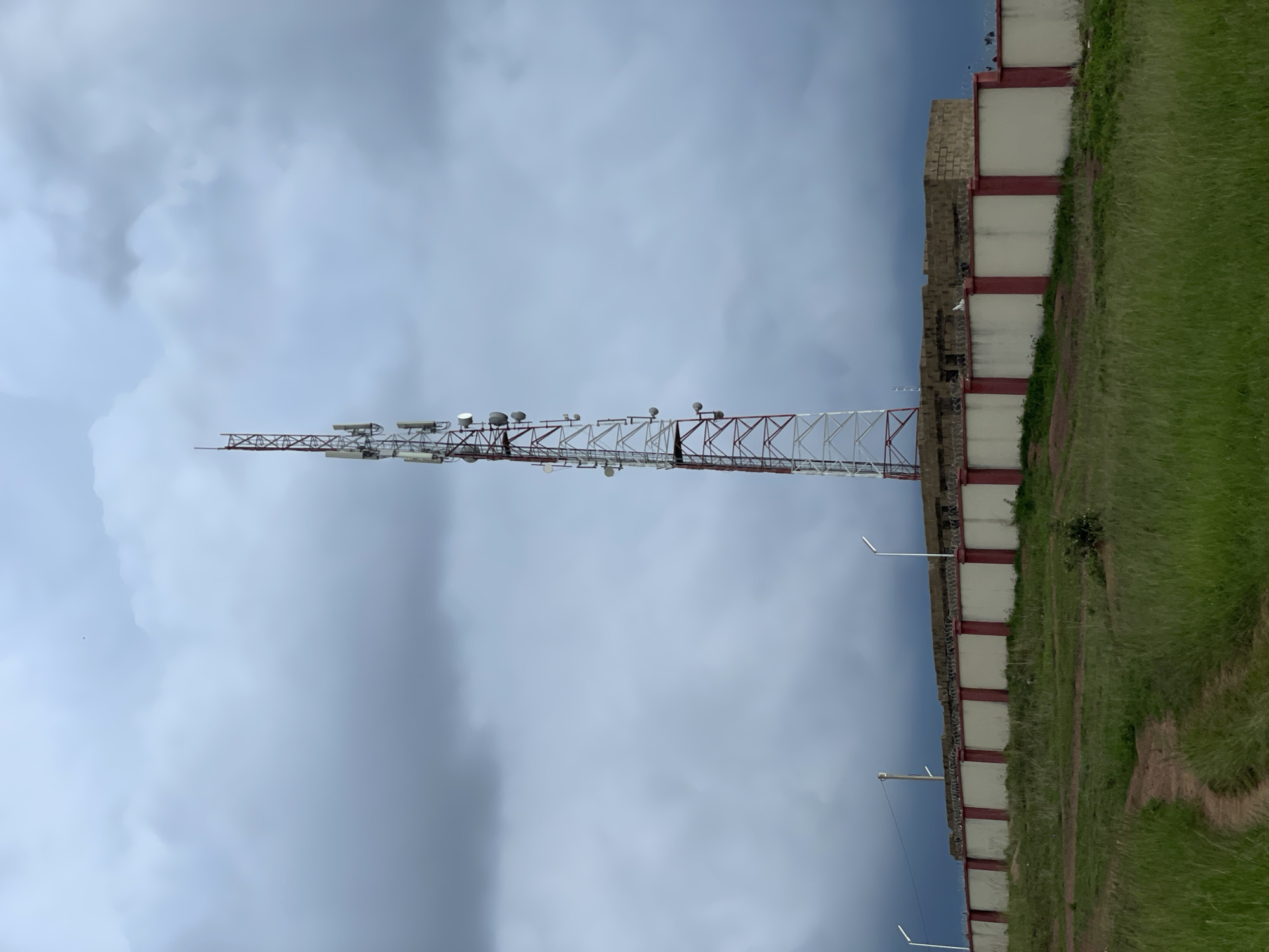 The image captures a telecommunications tower situated atop a walled enclosure in Durumi, Nigeria. The tower, a red and white lattice structure, is adorned with numerous antennas and equipment. The enclosure is characterized by a white wall with red pillars, topped with barbed wire. Beyond the wall, a building constructed of light-colored bricks is visible, and further back, a dark, cloudy sky looms. The foreground features a grassy area with a dirt path winding through it. The scene is devoid of any people or observable activity, presenting a static, environmental portrait.