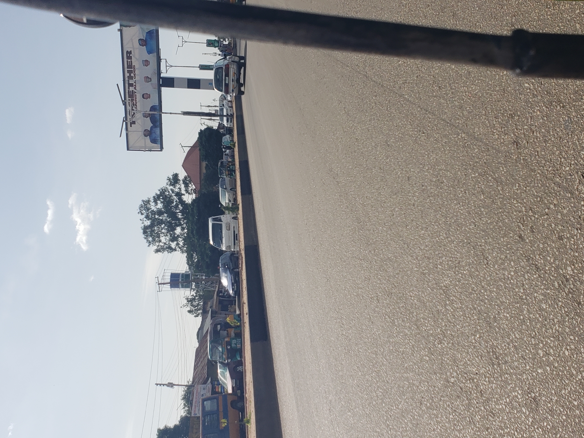 A street scene in Jos, Nigeria, is depicted on a bright, clear day. In the foreground, a paved asphalt road occupies the majority of the frame. A dark, vertical pole, likely part of a vehicle or structure, intrudes on the right side of the image.
Beyond the road, a raised concrete barrier separates the traffic lanes from a sidewalk or verge. Along this verge, several vehicles are parked or in motion. On the left, a blue and yellow bus is visible, followed by a car and then a tricycle, commonly known as an "Aja" or "Keke Napep," with a yellow canopy. Further down the road, a dark blue car, a white utility van, and a silver minivan are present.
A large billboard with a political advertisement stands prominently against the sky. The billboard features the word "TOGETHER" in large font, with smaller text below it reading "AGAINST ALL ODDS." Five men in traditional attire are pictured on the billboard.
The background consists of trees, buildings with corrugated iron roofs, and utility poles. A tall, dark vertical structure, possibly a water tower or a radio mast, is situated behind the billboard, with electrical wires connecting to it. The sky is a pale blue with scattered white clouds. The overall impression is of a busy urban street environment in Nigeria.