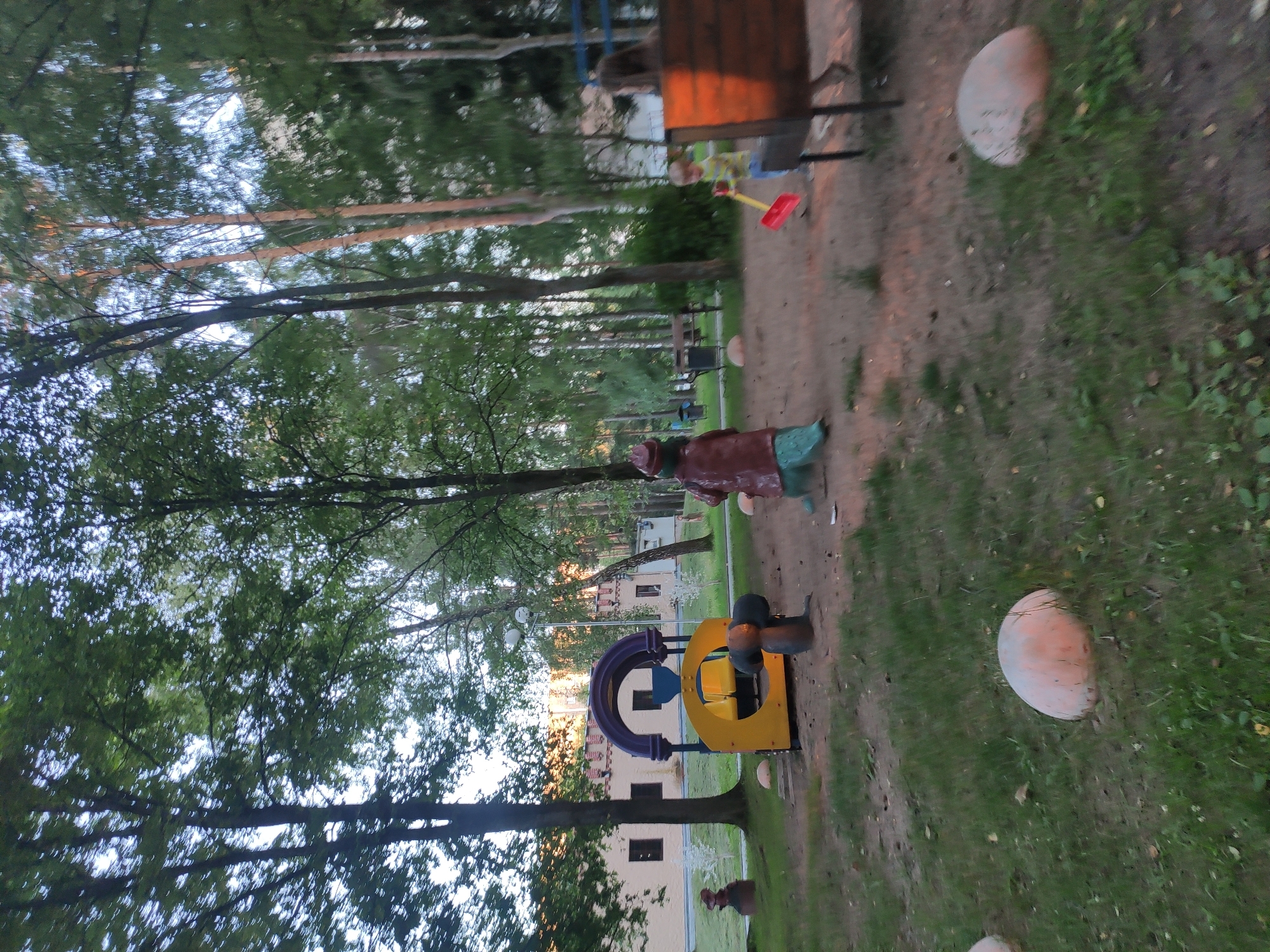 The image depicts a playground in Navapolatsk, Belarus. In the foreground, a child with blonde hair is playing with a red shovel and broom. To the left, there is a statue of a figure in a brown coat and a green garment, positioned in front of a yellow and purple playground structure that resembles a train. A building with a beige facade and arched windows is visible in the background. The playground is surrounded by trees and has a sandy ground. Several large, smooth, orange-pinkish rocks are scattered on the grassy areas of the playground.