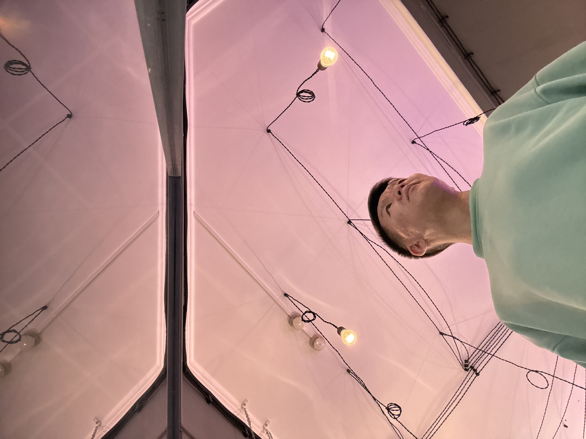 The image shows a man looking up at a tent-like structure with lights strung inside. The structure is made of white material stretched over a frame, and the lights are suspended by black wires. The man is wearing a light green sweater. The perspective is from below, looking up at an angle. There's a reflective surface, possibly a mirror or glass panel, on the left side of the image, reflecting the tent structure and lights. The ceiling above the tent appears to be dark grey or black. There is no information on the location of this image, the city of Moscow, Russia.