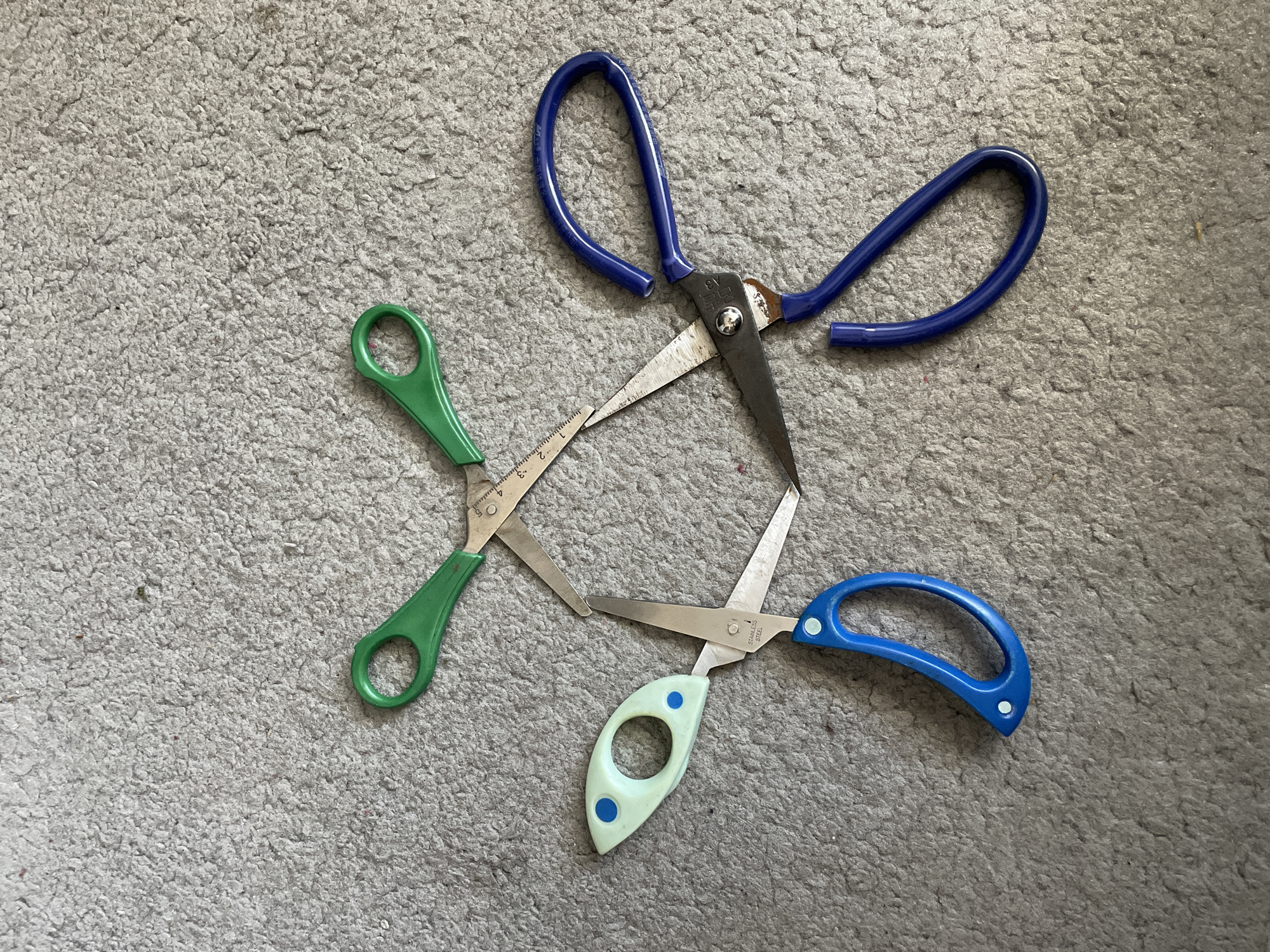 The image depicts three pairs of scissors arranged in a haphazard configuration on a gray carpet. The scissors have handles of blue, green, and light green with blue spots. The blades of the scissors intersect at their pivot points. The blue-handled scissors appear to have a broken handle. The environment suggests an indoor setting. There is no indication that the image was taken in Asenovgrad, Bulgaria, or any other specific location. There are no people present in the image.