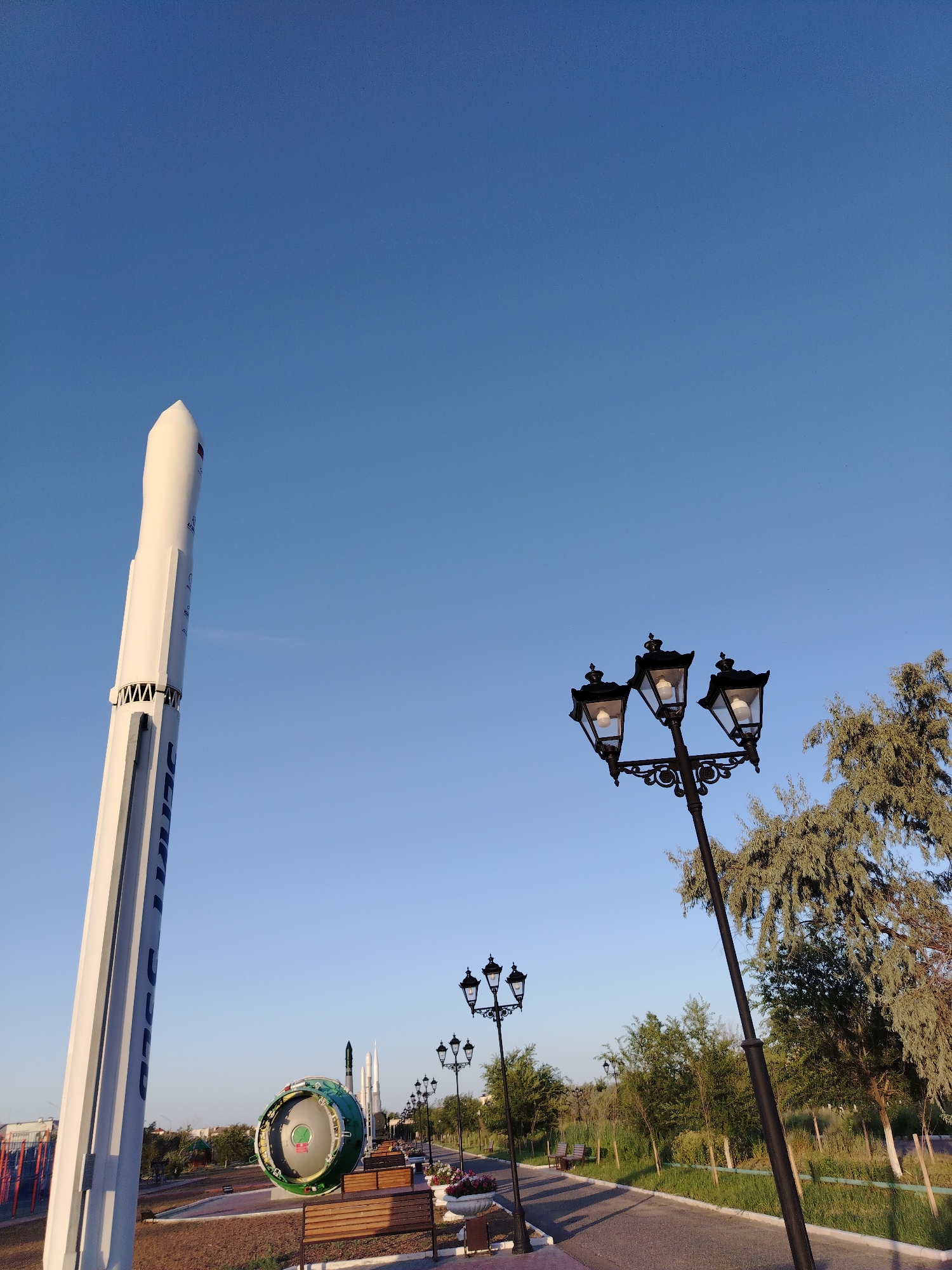 This image depicts a section of the Baikonur Cosmodrome park in Baikonur, Kazakhstan, under a clear blue sky. In the foreground and to the left, a tall, white rocket monument stands, its base partially obscured by the camera's angle. To its right, a pathway lined with black, ornate streetlights, each with three lamps, curves into the distance. A large, circular object, possibly a spacecraft capsule or part of a rocket engine, painted in green and silver, is prominently displayed on the left side of the pathway, further back from the rocket monument. Behind these exhibits, a neatly manicured park area features benches, trees, and flowerbeds. The overall scene conveys a sense of public space dedicated to space exploration heritage. There are no people visible in the image.