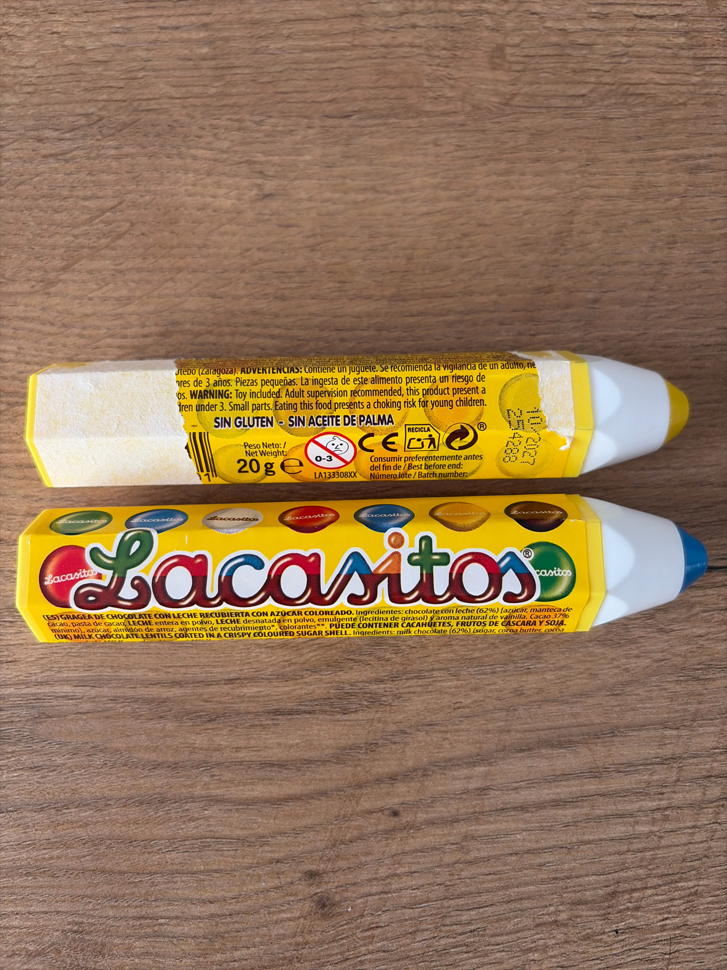 Two yellow, pencil-shaped candy packages are placed on a wooden surface. The top package is partially unwrapped, revealing text and a barcode. The bottom package is fully intact and displays the brand "Lacasitos" prominently. The text on both packages is in Spanish and English, indicating the product is chocolate with a colored sugar shell and includes a warning about small parts for children. The wooden surface appears to be a table with a visible grain. There are no people or animals present in the image. The scene is static and appears to be a product shot. The location of Oxentea, Moldova is not discernible from the image.
