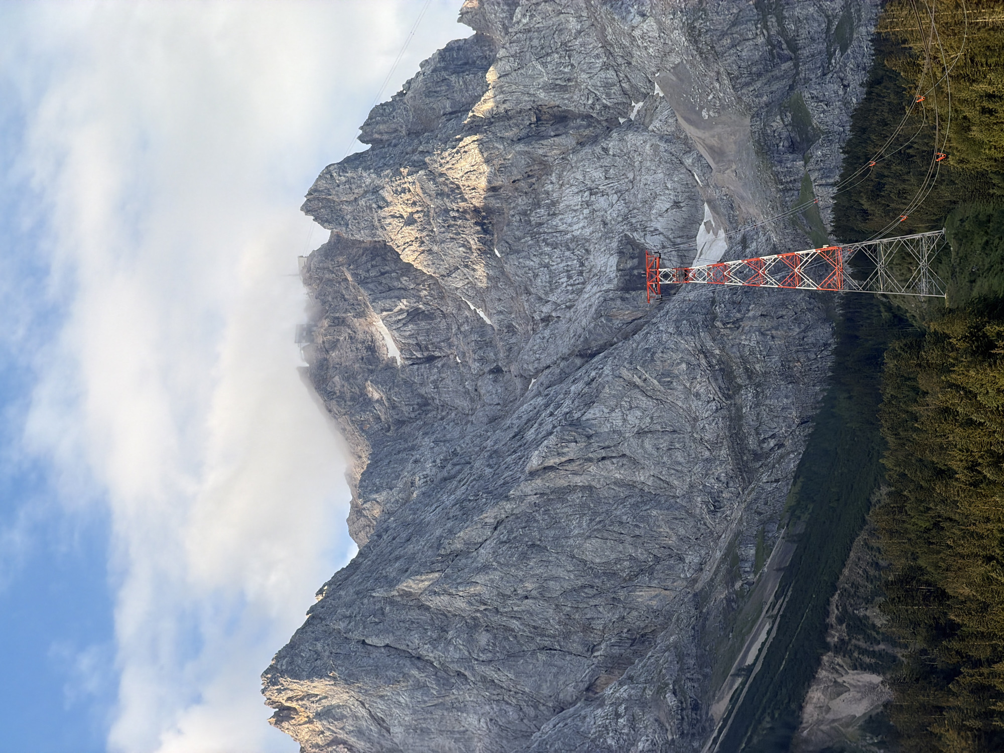 The image captures a majestic mountain landscape in Grainau, Germany. Dominating the scene is a colossal, rugged mountain with a steep, rocky face. The upper reaches of the mountain are partially obscured by wisps of white clouds. A prominent, red and white lattice tower stands in the foreground, supporting cables that extend towards the mountain. The lower slopes of the mountain are covered in a dense forest of dark green coniferous trees. The sky above is a clear blue, dotted with scattered white clouds. The overall impression is one of a dramatic natural setting with a man-made structure integrated into the environment.