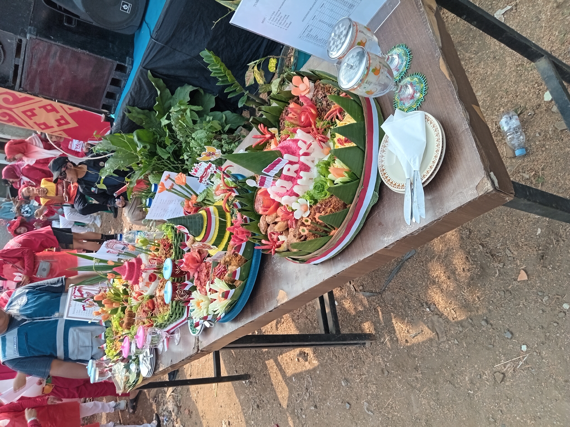 The image depicts an outdoor event, possibly a celebration, on a dirt and gravel ground. The scene features a long wooden table laden with food, specifically elaborate "Tumpeng" creations, which are cone-shaped rice dishes often adorned with vegetables and meat. Several people are present, some wearing red and white attire, suggesting a patriotic theme or a specific event. One person in the foreground wears a blue vest over a dark shirt. In the background, there are large speakers and a stage draped in black fabric. A sign with Indonesian text is visible on the table. The overall impression is of a festive gathering with a focus on food presentation. The context of Depok, Indonesia, suggests the cultural significance of the Tumpeng and the likely presence of Indonesian people participating in the event.