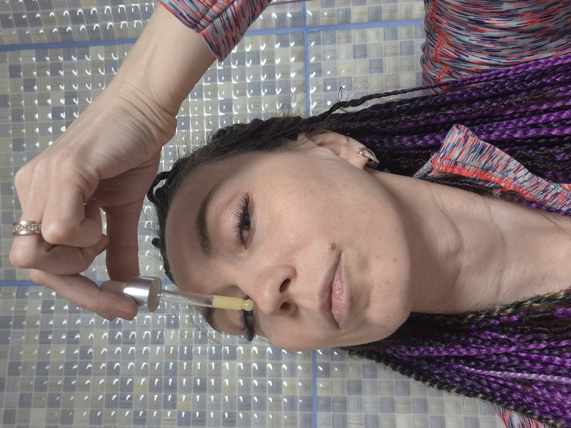 A woman with purple braided hair is applying a yellowish liquid from a pipette near her nose. She is wearing rings on her fingers. The background is a wall tiled with square tiles in shades of grey and white. The location is Chaikovsky, Russia.