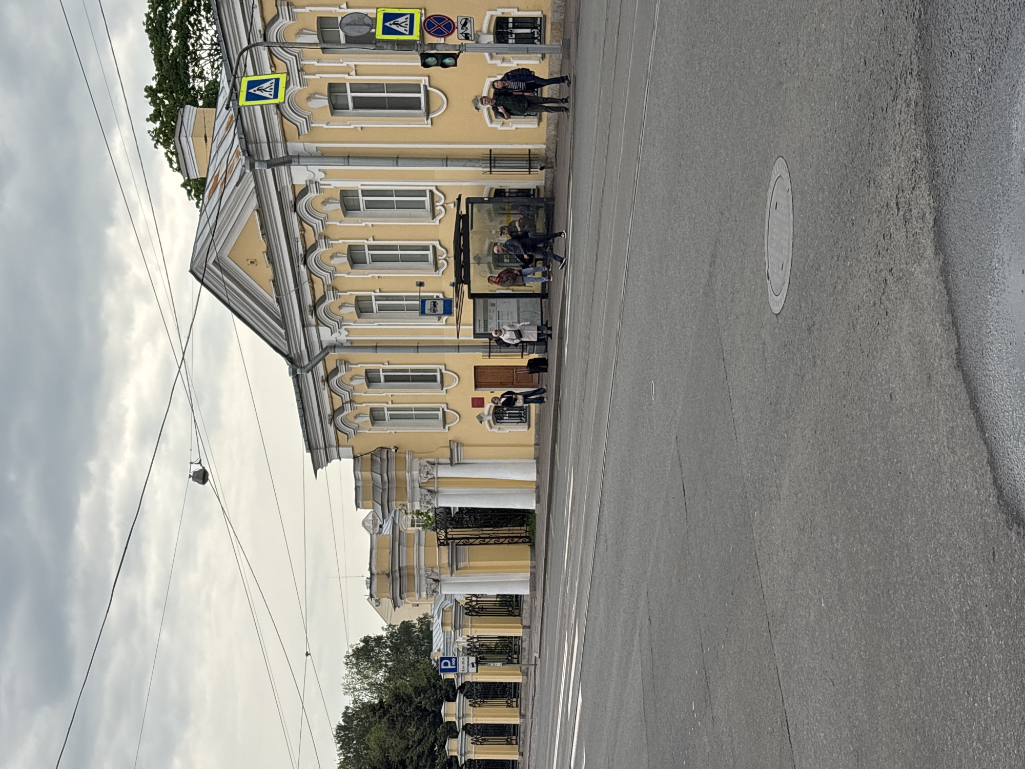 The image depicts a street scene in Saint Petersburg, Russia. A yellow building with white columns and trim occupies the left side of the frame. Pedestrian crossing signs and a traffic light are visible near the building. People are waiting at a bus stop next to the building. Some individuals are standing on the sidewalk adjacent to the building. A section of the road is visible on the right.