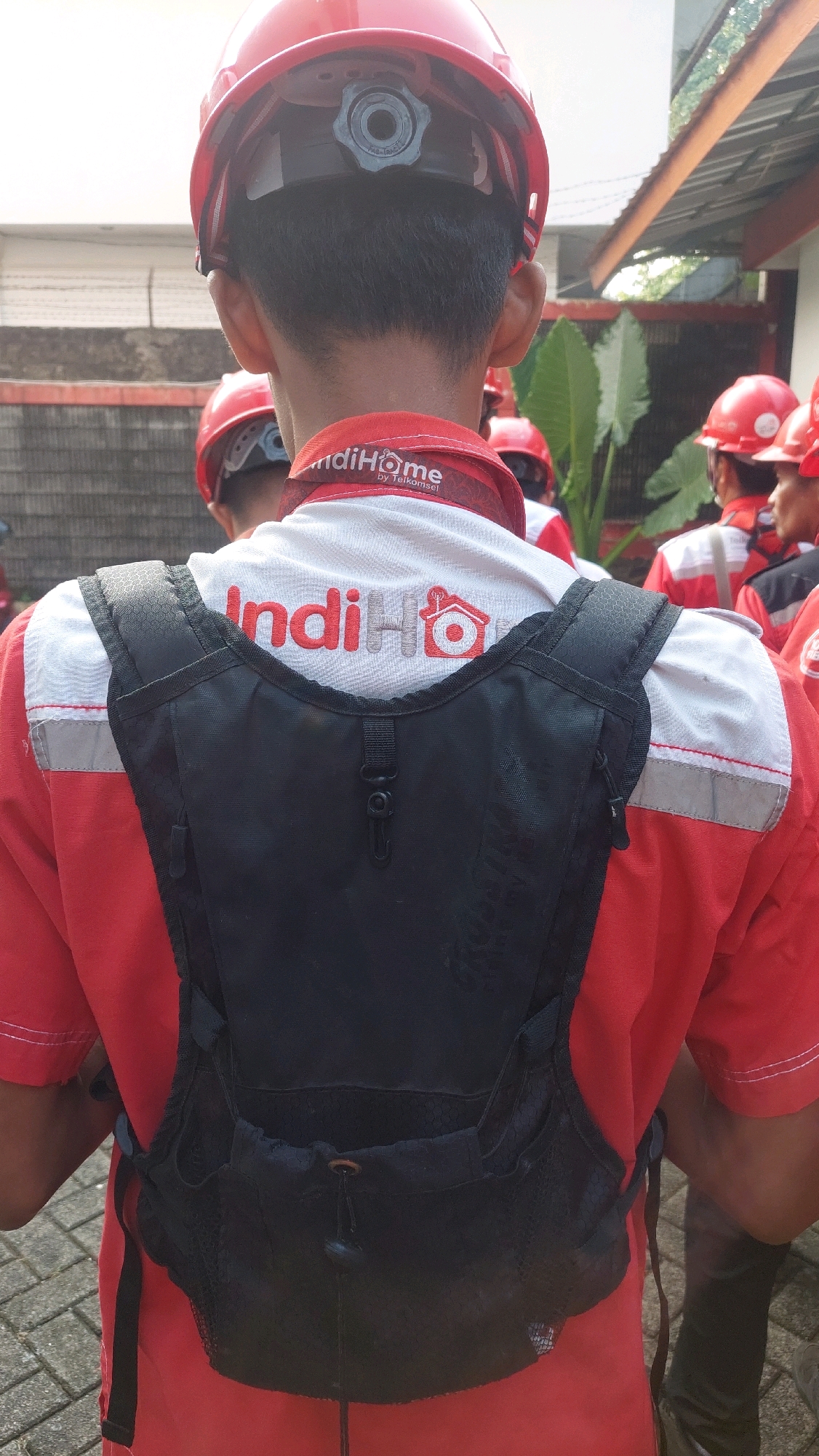 The image shows the back of a person wearing a red and white uniform, a red hard hat, and a black backpack. The uniform has "IndiHome" written in red letters across the back, with a house logo. The person is standing outdoors on a paved surface, possibly a sidewalk or road. In the background, other individuals wearing similar uniforms and red hard hats are visible, along with some foliage and buildings. The setting appears to be a semi-urban environment in Cibinong, Indonesia, likely associated with a telecommunications company.