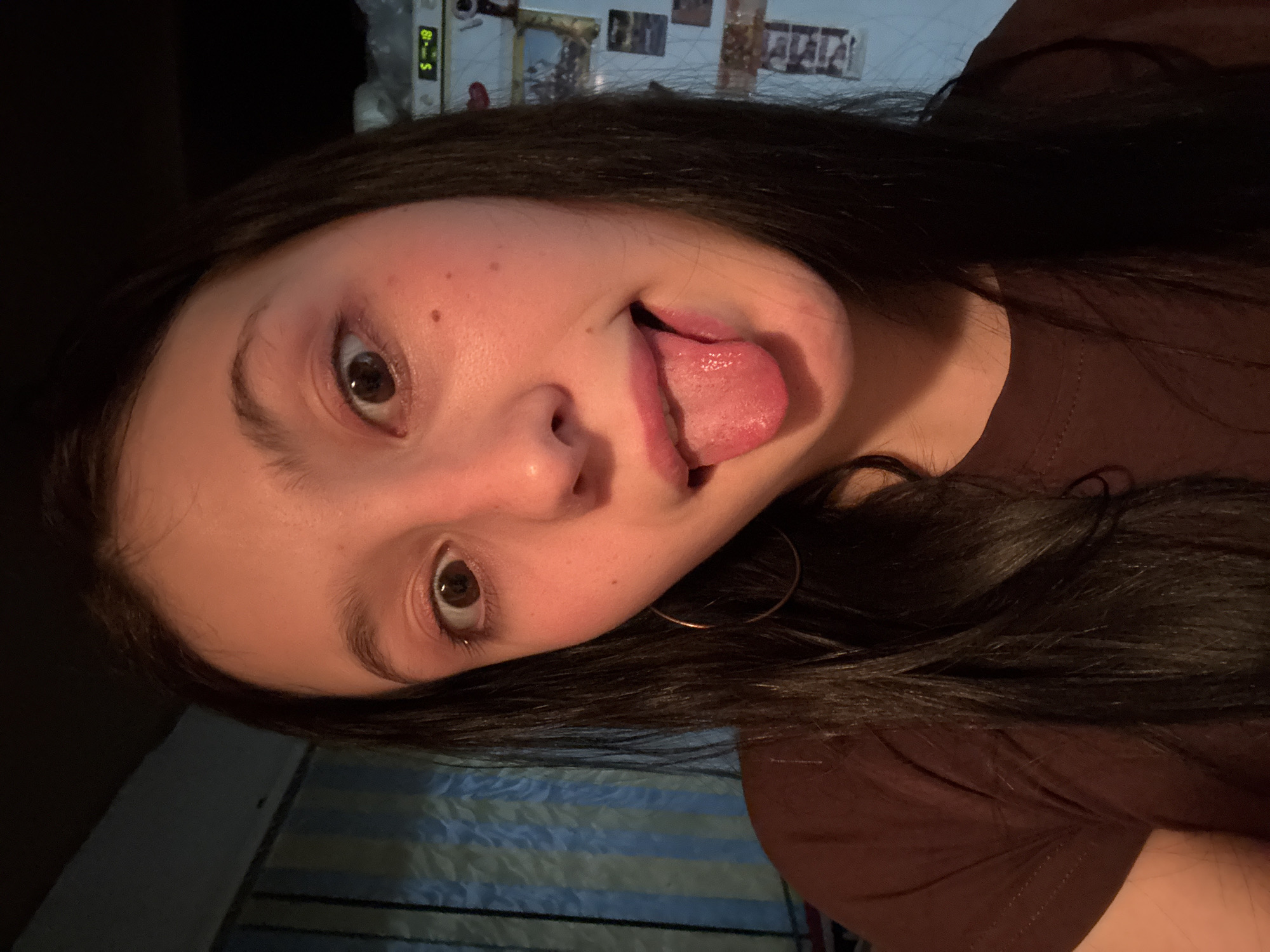 The media file presents an angled portrait of a person, likely a young woman, with long dark hair and brown eyes. The subject is wearing a dark brown t-shirt or top and is captured in a playful pose with their tongue slightly sticking out to the side. A large hoop earring is visible on the right side of their face.
The scene is indoors, dimly lit, suggesting low-light conditions, possibly evening or night. In the upper background, a digital clock with green illumination clearly displays "5 - 18". Faint outlines of pictures or frames are visible on a wall behind the subject. In the lower part of the image, a striped fabric, potentially a bedsheet or blanket, in light blue and white/beige patterns is visible. The lighting casts a warm tone on the person's skin. The location context for this media file is Фоки, Russia.