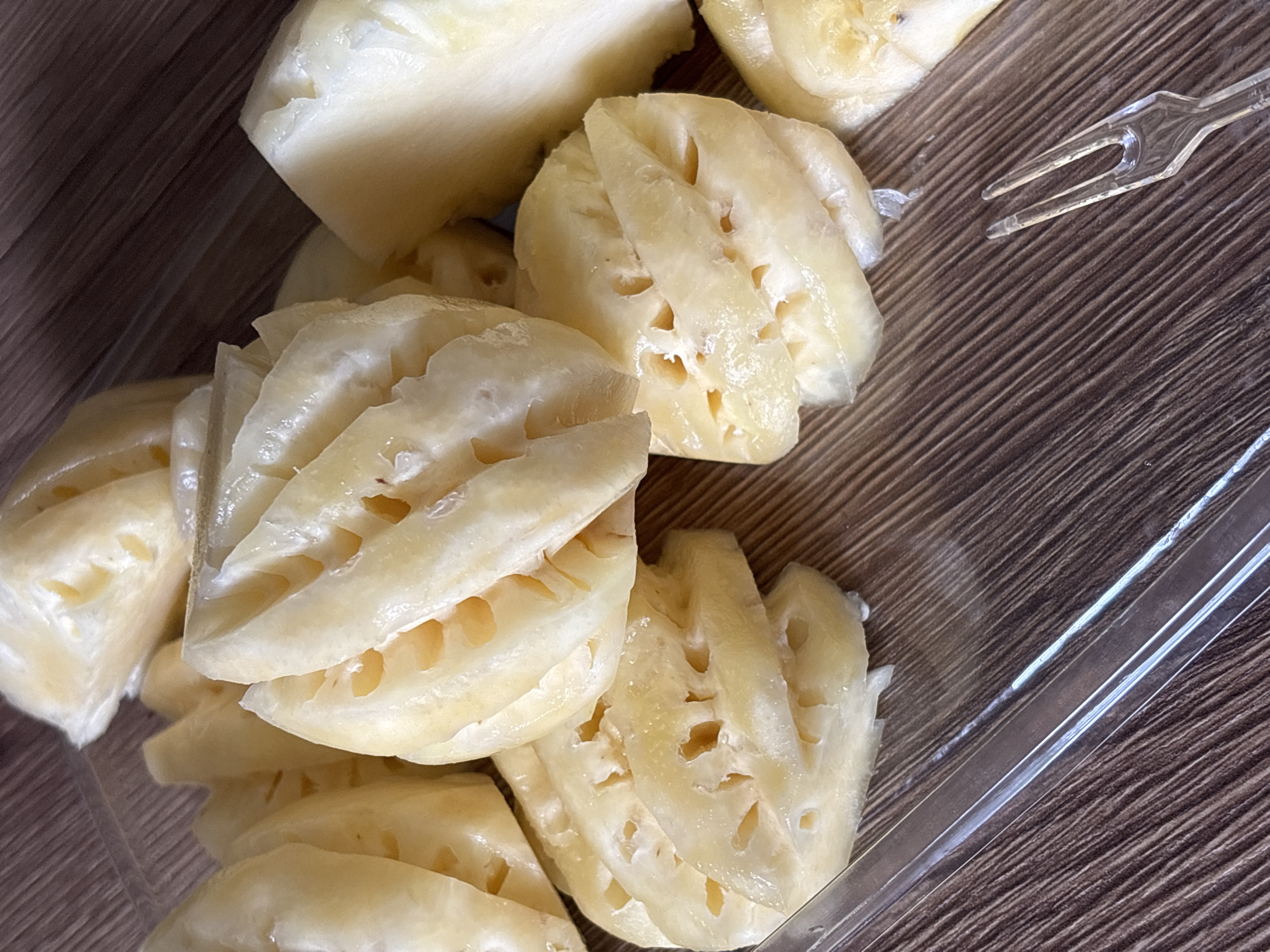 The image shows a close-up view of cut lotus root in a plastic container on a wood-grain surface. A clear plastic fork and lid are also present. The scene is likely indoors, given the controlled environment. The specific location in Choeng Thale, Thailand, is not discernible from the image alone. No people, actions, or interactions are visible.