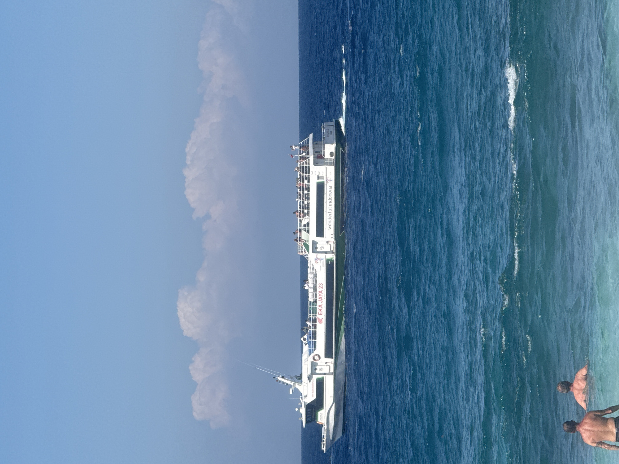 A ferry boat, named "EKA JAYA 23", travels across a bright blue ocean under a clear sky with scattered white clouds. The ferry has a white hull with a green stripe and the text "WONDERFUL INDONESIA" printed on its side. Several passengers are visible on the upper deck of the ferry, enjoying the ride. In the foreground, two men are partially submerged in the ocean, facing away from the camera. The water is calm with gentle waves. The scene appears to be a tourist location in Manggis, Indonesia, possibly a coastal area known for its ferry services and beaches.
