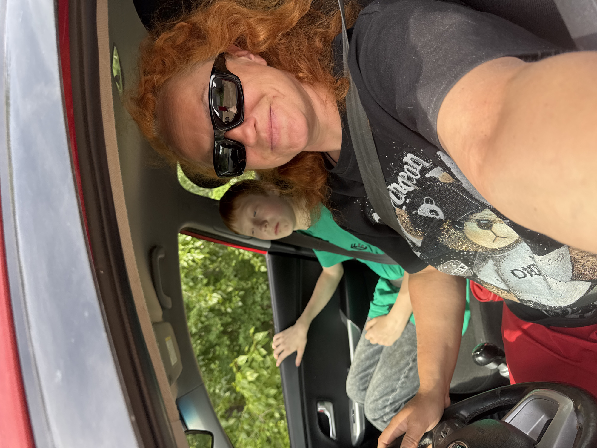 This image captures a selfie taken inside a car, featuring a woman in the driver's seat and a child in the back. The woman, with red, curly hair and dark rectangular sunglasses, looks directly at the camera with a slight smile. She is wearing a black t-shirt adorned with a sparkling design that includes what appears to be a teddy bear's head and partially visible text. She is secured by a seatbelt, and her hands are on the steering wheel, which features a clearly visible "FIAT" logo.
In the back seat, to the woman's left, a child with reddish-brown hair is visible. He is wearing a green t-shirt and grey pants, looking generally forward or downwards, with his hand resting on the car's door frame.
The car's interior is visible, including the seatbelt, steering wheel controls, and parts of the dashboard and door. Through the car windows, lush green foliage, indicative of trees or bushes, suggests an outdoor setting during bright daylight. The exterior of the car appears to be red in color, visible on the door frame. The scene conveys a casual, perhaps leisurely moment during the day. The location context for this media file is Chaikovsky, Russia.
