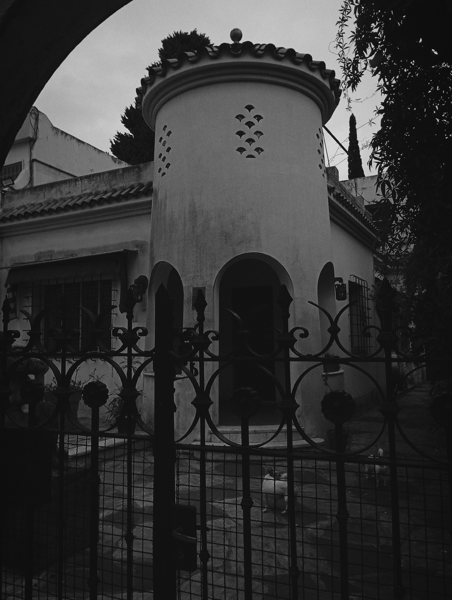 A black and white photograph depicts a house with a tower behind a wrought iron gate. The house has a rounded tower on its front facade, featuring decorative cutouts. An arched entryway leads into the house. Windows are visible on either side of the entrance. A second story can be seen behind the main structure, partially obscured by trees. The wrought iron gate, in the foreground, is partially obstructing the view of the house. The city of Buenos Aires, Argentina, cannot be determined from the image.