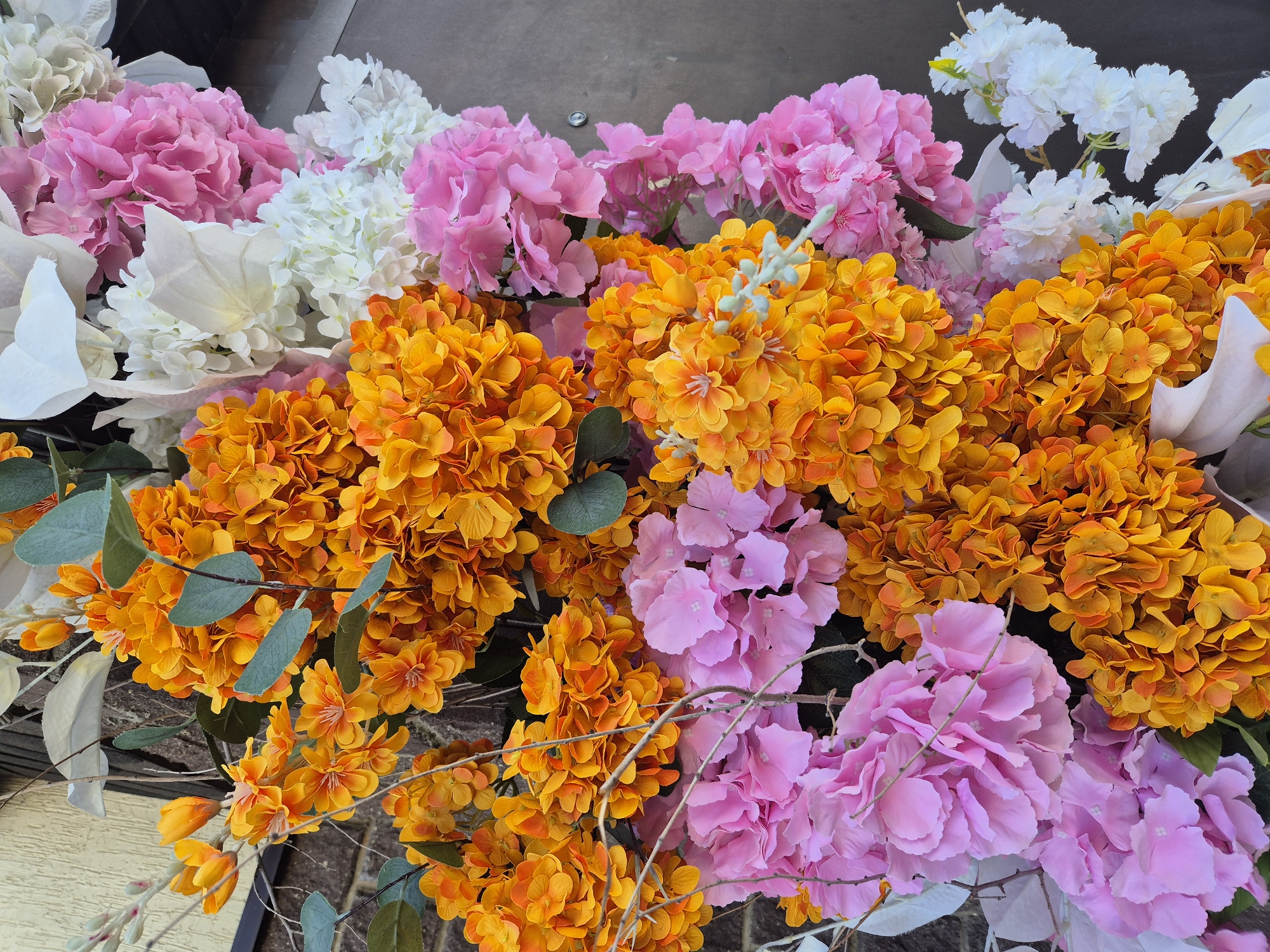 This is a close-up, overhead shot of a vibrant floral arrangement, possibly a wedding arch or a decorative installation. The flowers are predominantly artificial and feature a mix of colors, including bright orange, soft pink, and white. The orange flowers are clustered densely, resembling hydrangeas or marigolds, while the pink flowers are also clustered and appear to be hydrangeas. White flowers, some with delicate petals and others with a more solid form, are interspersed throughout the arrangement. Some green artificial leaves and thin, dry branches are also visible, adding texture and depth. The background consists of a textured, light-colored wall on the left and a dark, neutral surface on the right. There are no people or animals visible in the image, and no specific urban features or landmarks of Kokshetau, Kazakhstan are identifiable. The scene is static and solely focuses on the floral display.