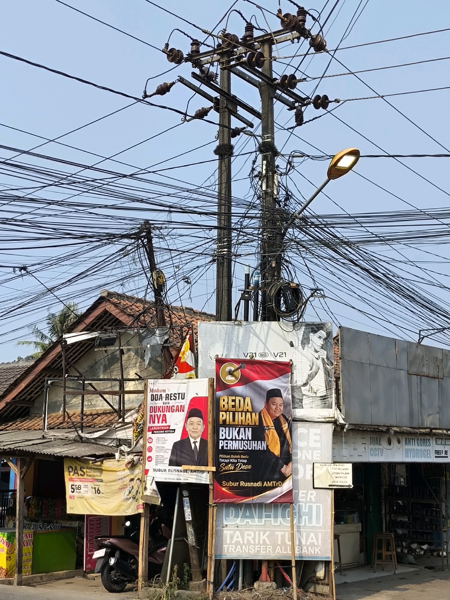 The image depicts a street scene in Lubangbuaya, Indonesia, characterized by an overhead tangle of electrical wires. A prominent utility pole dominates the frame, supporting numerous wires that crisscross the sky. A street lamp is visible on the right side of the pole. In the foreground, several advertisements are displayed. One large red and white banner features a man in traditional attire and text in Indonesian, likely a political or campaign poster. Another yellow banner advertises a mobile data plan with "PAS" and pricing information. To the right, a shop storefront is visible, with signs indicating it sells items such as "ANTI GORES," "HYDROGEL," and "FREE FIRE." A motorcycle is parked on the left side of the image. The environment appears to be an urban or semi-urban setting with modest buildings and a clear blue sky.