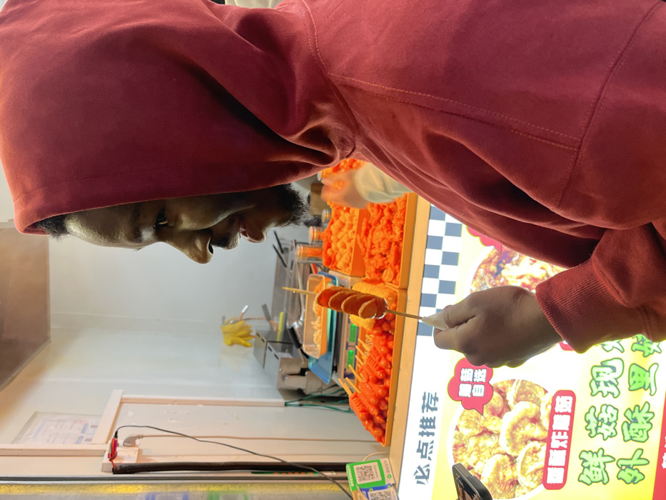 The image depicts a male individual, observed in profile facing right, holding a skewer of food at a street food stall in Jiangjin, China. The man, with a dark complexion and a short beard, wears a red hooded sweatshirt with the hood pulled up. He holds a wooden skewer with his right hand, displaying five cylindrical, sliced, reddish-brown food items, likely fried sausages, coated in a light yellow batter. His mouth is slightly ajar, and he exhibits a subtle smile.
In the background, a brightly lit food display counter features multiple orange rectangular trays filled with various items, including numerous skewered sausages identical to the one he holds, and piles of smaller, round, orange-colored fried snacks. A prominent sign in the foreground displays Chinese characters, including "必点推荐" (Bì diǎn tuījiàn - Must-order recommendations), alongside images of other food items like fried mushrooms or dumplings. To the left of the counter, two green QR codes are visible, indicating mobile payment options. The background also shows a plain white wall with a power outlet and wires, a metal counter with scales, and a yellow glove hanging. The environment is consistent with an illuminated indoor or covered food vendor stall.