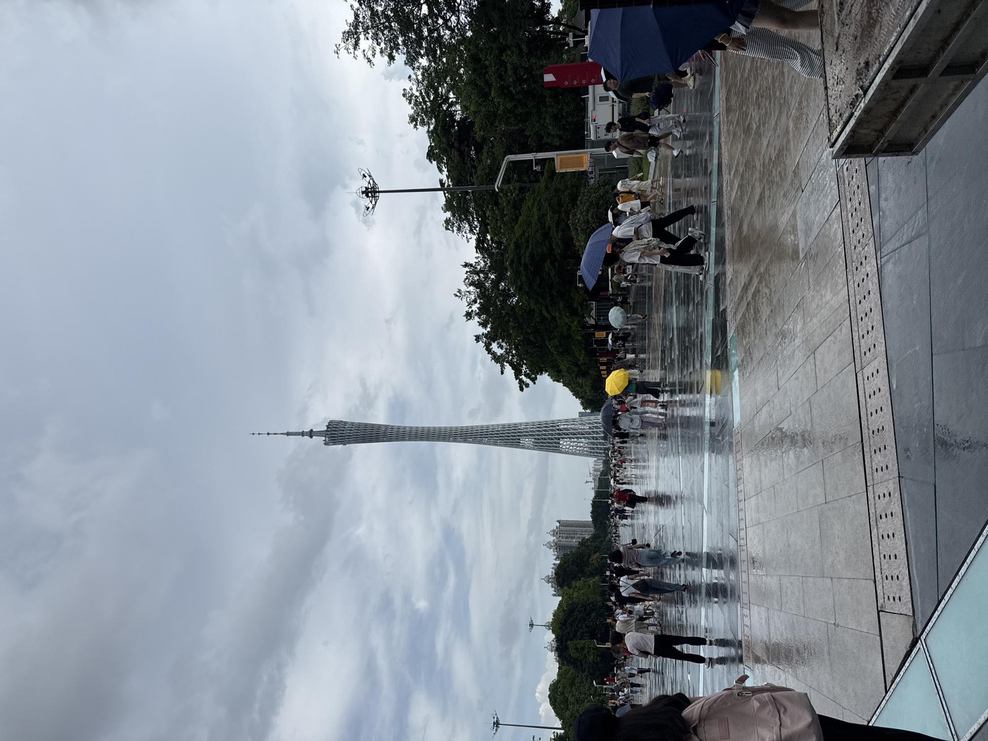 The image depicts a wide, wet, paved pedestrian area in Tianhe District, China, with numerous people walking, some holding umbrellas. A tall, slender, lattice-like tower dominates the background, its distinctive shape indicative of the Canton Tower. The ground is reflective due to recent rain, showing the shapes of people and the surrounding environment. Trees line the sides of the area, and modern buildings are visible in the distance. Streetlights and traffic signals are present, suggesting an urban setting. The sky is overcast with heavy clouds. People are dressed in casual attire, and the overall atmosphere suggests a busy public space during or after rainfall.