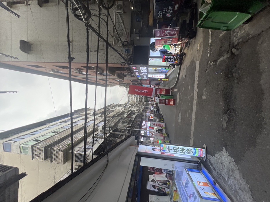 The image depicts a narrow commercial street or alleyway in Jiangjin, China, under an overcast sky. Two multi-story buildings flank the asphalt street, which shows signs of wear including cracks and potholes.
On the left side, a ground-level storefront displays electronic devices in glass cases. A prominent, illuminated vertical sign in Chinese reads "手机维修" (Mobile Phone Repair). Above the storefront, the building features several stories with multiple balconies, windows, and visible air conditioning units, presenting an aged light-colored facade. Overhead, a dense network of black electrical and communication cables stretches across the street.
On the right side, in the immediate foreground, a large green wheeled trash bin is partially visible. Further along the right, a series of storefronts display illuminated advertisements for electronics and mobile devices. A large red banner vertically displays the "HUAWEI" logo in white. Standing next to this banner is a person in a bright green uniform, facing away from the viewer. Parked near these shops are two scooters or motorcycles and a three-wheeled utility vehicle with Chinese characters on its side. The building on the right is also multi-story, with a reddish-brown facade that appears to transition to a lighter color further up, featuring windows and air conditioning units.
In the background, the street recedes between more buildings, and several indistinct vehicles are visible. A yellow safety cone stands on the left side of the street further down. The overall scene suggests an active urban commercial area.