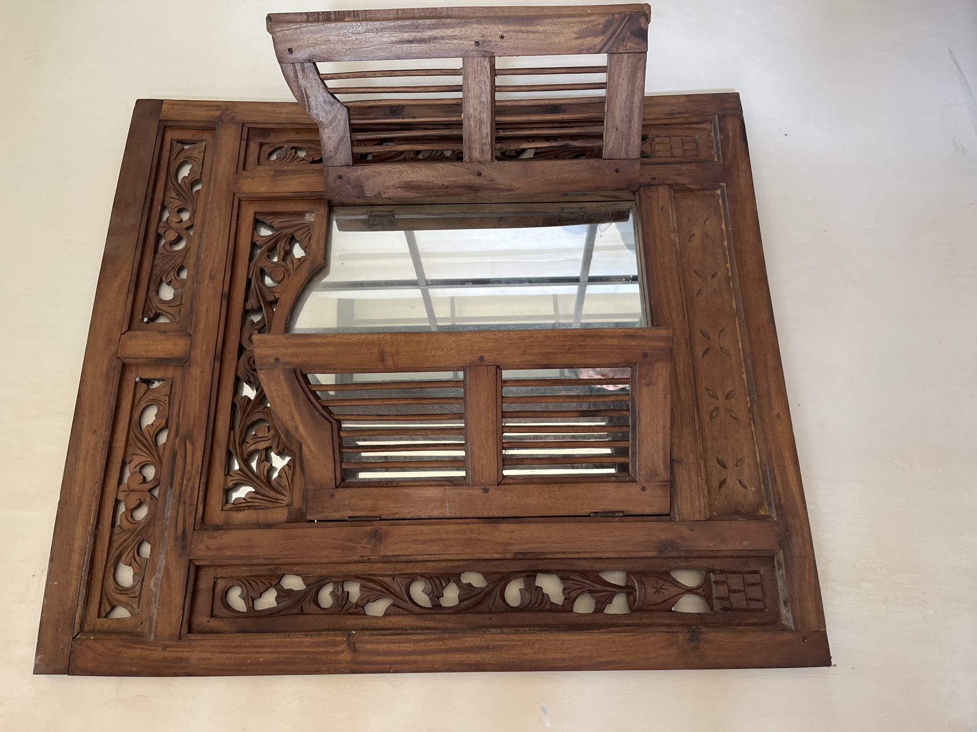 An ornate wooden window frame, likely made of teak, is mounted on a textured, off-white wall. The frame features intricate carvings of floral motifs and geometric patterns. The window itself has two panels, both with vertical wooden slats. The left panel is closed, while the right panel is open outwards at a roughly 90-degree angle. The interior of the window reveals a reflective surface, likely glass, showing a blurred reflection of a person and what appears to be a white curtain. The window is located in the city of Sidoarjo, Indonesia, a region known for its skilled woodcarving traditions, particularly from the nearby Javanese culture. The overall impression is one of traditional craftsmanship and an aesthetic that evokes a sense of history and cultural heritage.