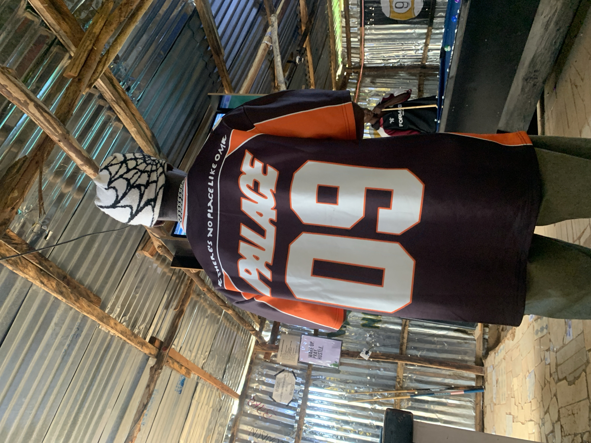 The image depicts an indoor scene, likely a recreational area, possibly a bar or a pool hall, in Durumi, Nigeria. The foreground is dominated by a person from behind, wearing a dark jersey with "PALACE" and the number "09" emblazoned in white and orange. The jersey also has text on the upper back reading "THERE'S NO PLACE LIKE OMR". The person wears a white beanie with a black spiderweb pattern and a thick silver chain necklace.
The background reveals a rustic, open-air structure with a corrugated metal roof supported by rough-hewn wooden beams and poles. The walls appear to be made of corrugated metal sheeting. Various signs are visible on the wall, including one with "NO CREDIT" and a phone number, and another with motivational text "WAKE UP. PRAY. HUSTLE." In the mid-ground, a pool table with several colored balls is visible. Another person stands near the pool table, holding a pool cue. The floor is a mosaic of irregular stone tiles. The overall impression is a casual, somewhat dilapidated yet functional establishment.