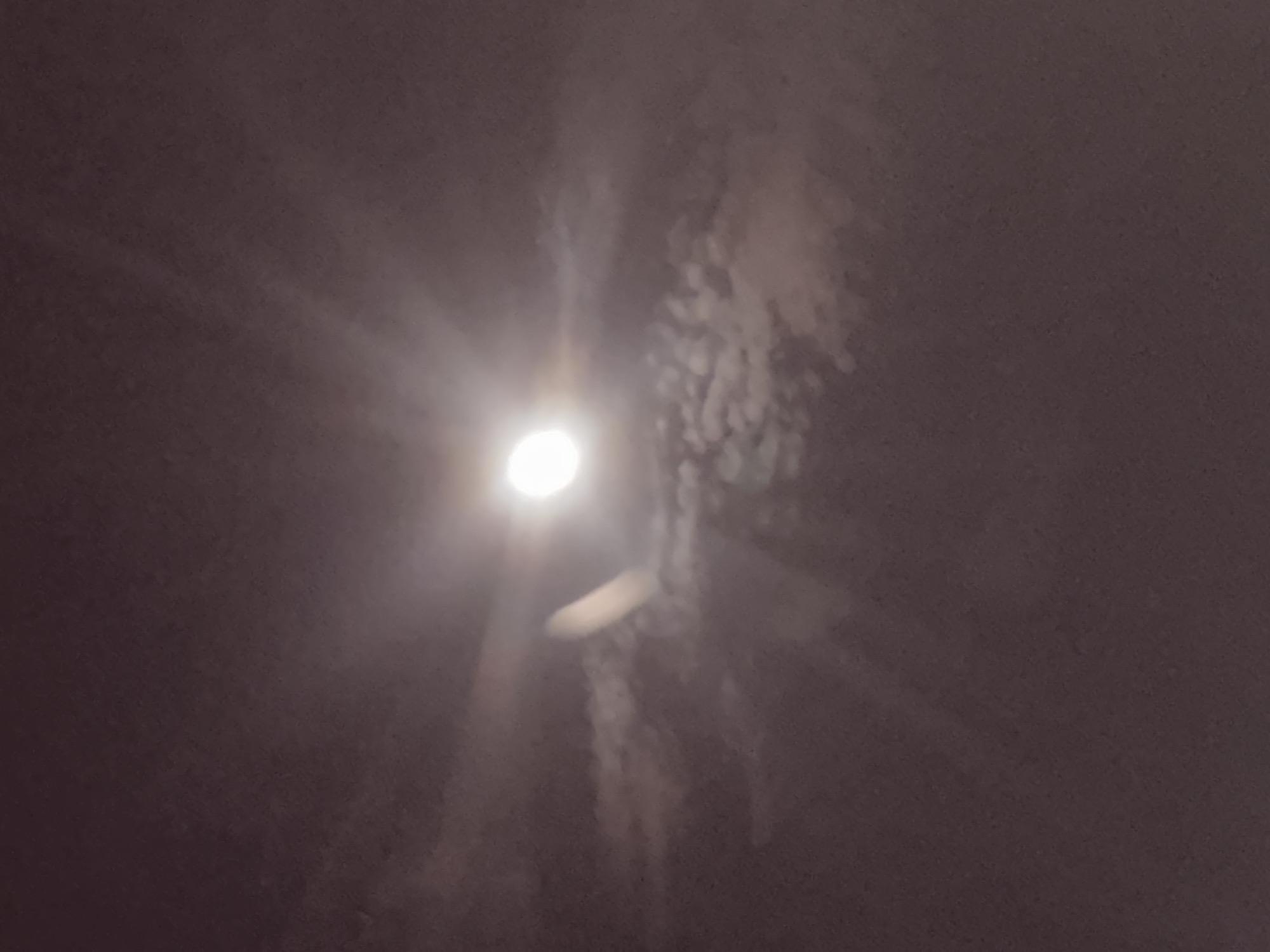 The image depicts a night scene, possibly outdoors, with a prominent bright light source, likely the moon or a strong artificial light, at the center-left. This light casts rays and illuminates a hazy or cloudy atmosphere, creating a diffused glow. To the right of the central light, there are streaks of lighter, textured material, possibly clouds or smoke, that appear to be illuminated from behind. There is also a blurred, elongated oval shape below and slightly to the right of the main light source. The overall impression is a dimly lit, possibly overcast night sky with a bright celestial body or light dominating the view. There is no visible text. The location context of Burgas, Bulgaria, suggests this could be a view of the night sky over the city or the Black Sea coast.
