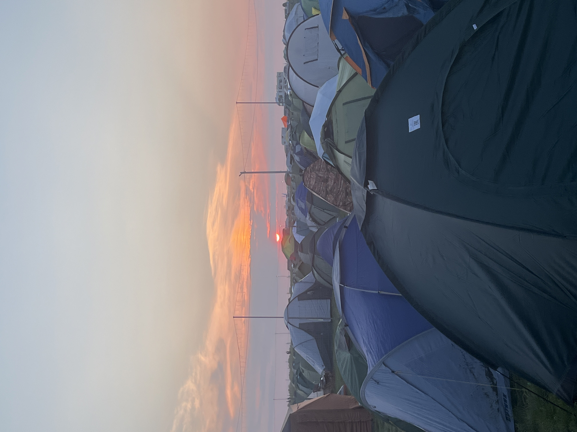 The image depicts a vast campsite filled with numerous tents under a soft, pastel-colored sky transitioning from pale blue to orange and pink, indicating either sunrise or sunset. The sun is visible as a small, bright orb near the horizon, partially obscured by clouds. A bridge-like structure with cables stretches across the middle ground, spanning a distance. In the background, several white recreational vehicles and other structures are visible. The foreground is dominated by a dense cluster of tents of various colors, including dark green, blue, and gray. Some tents are partially visible, creating a layered effect. The ground appears to be covered in grass. No people are clearly discernible in the image, and the scene is static, suggesting a time of rest or quiet. The overall atmosphere is peaceful and serene. The location is identified as Surakovo, Russia.