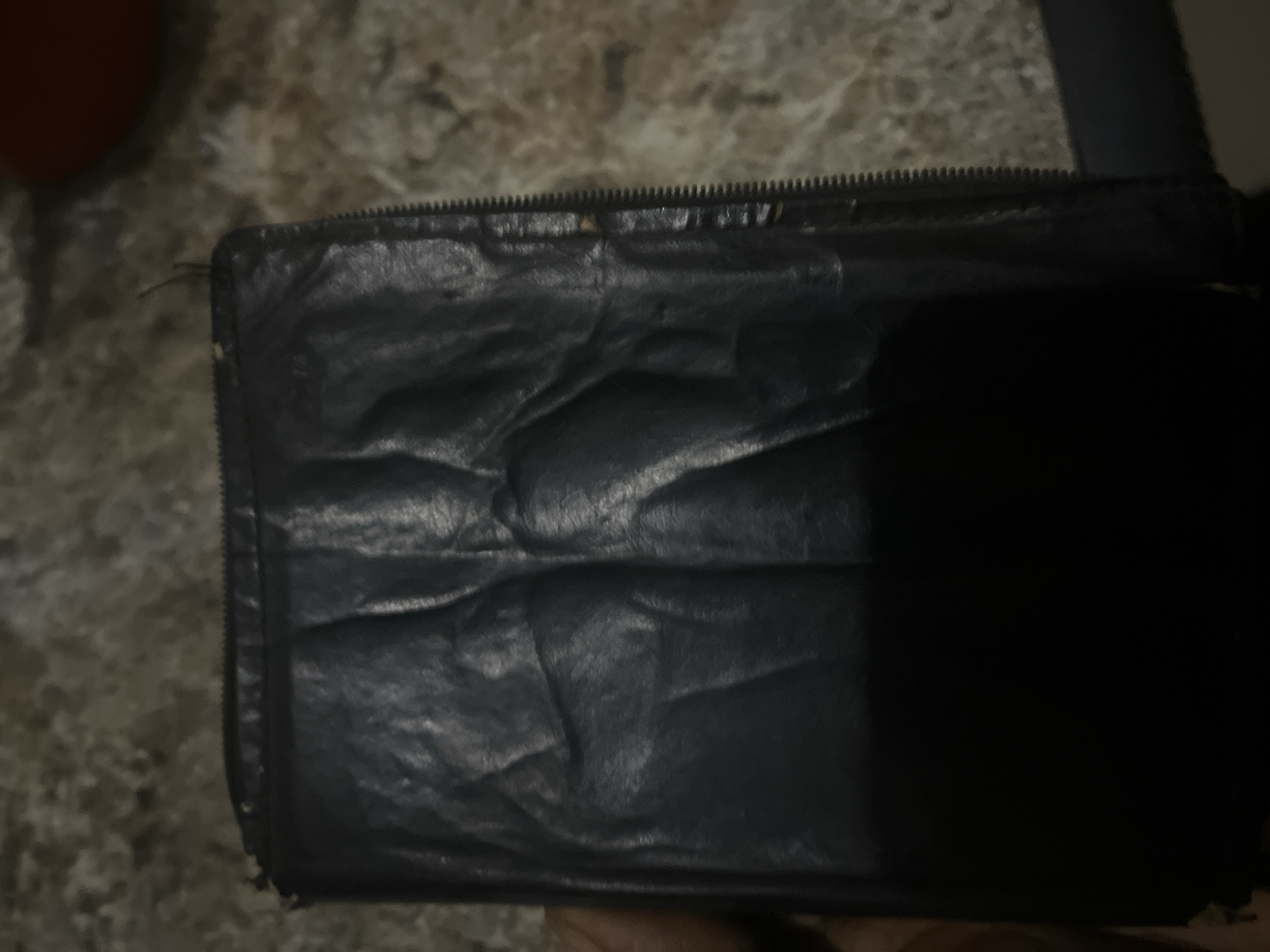 A close-up, overhead shot captures a dark-colored, wrinkled leather object, possibly a wallet or small bag, lying on a textured, light-brown surface. The object has a zipper closure visible along its right side. The lighting is dim, casting shadows and obscuring finer details of the object's texture and any potential contents. There are no people visible in the image, and the context of Jalingo, Nigeria, cannot be determined from the visual information provided.