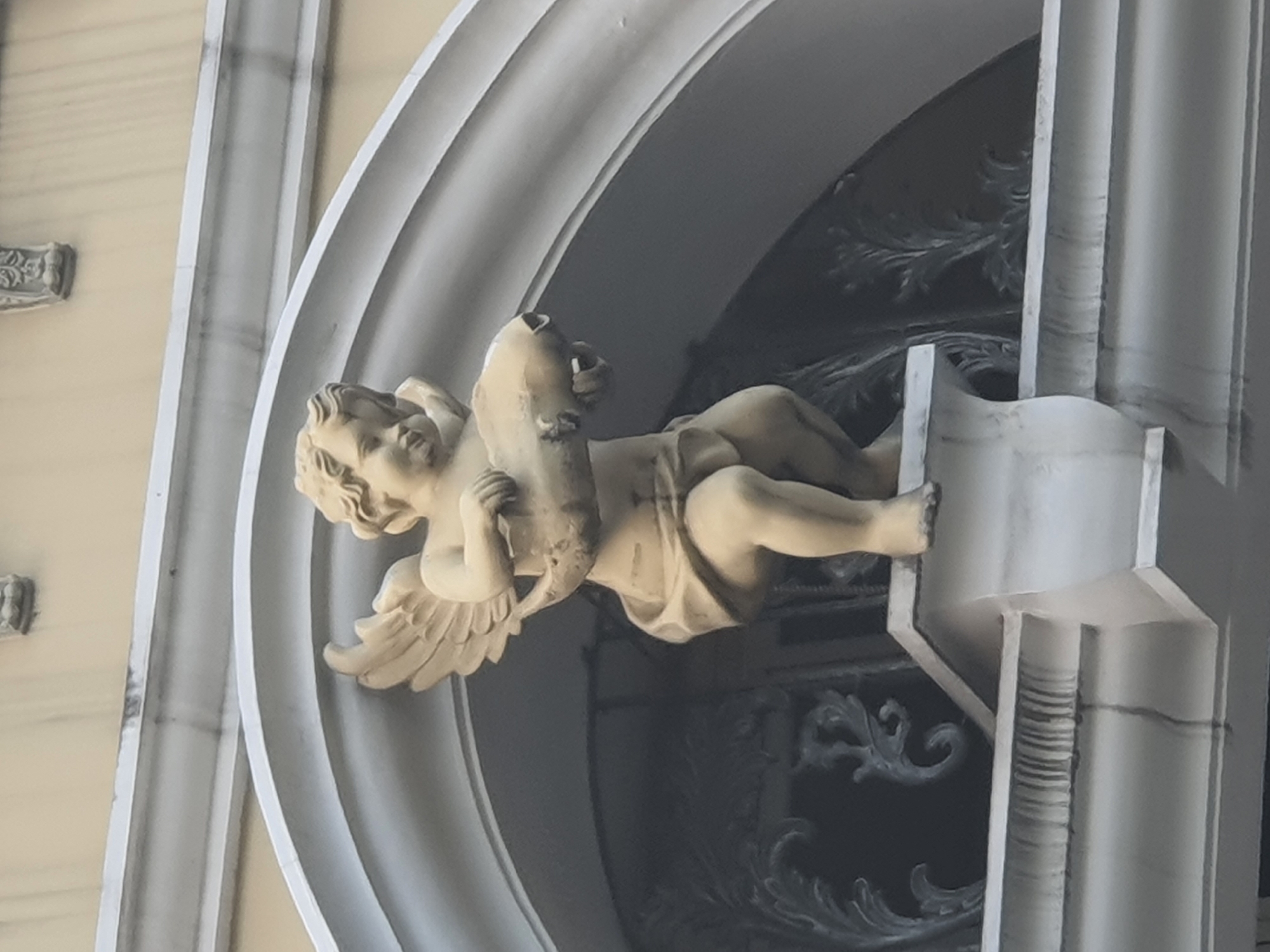 A light-colored stone statue of a cherub is positioned on a white architectural ledge. The cherub, depicted with wings, is holding a large, elongated, light-colored object that appears to be a fish. The statue is situated beneath an arched structure, possibly part of a building's facade. The background shows a wall with subtle decorative elements and further architectural details. The statue is located in front of a building in West Jakarta, Indonesia.