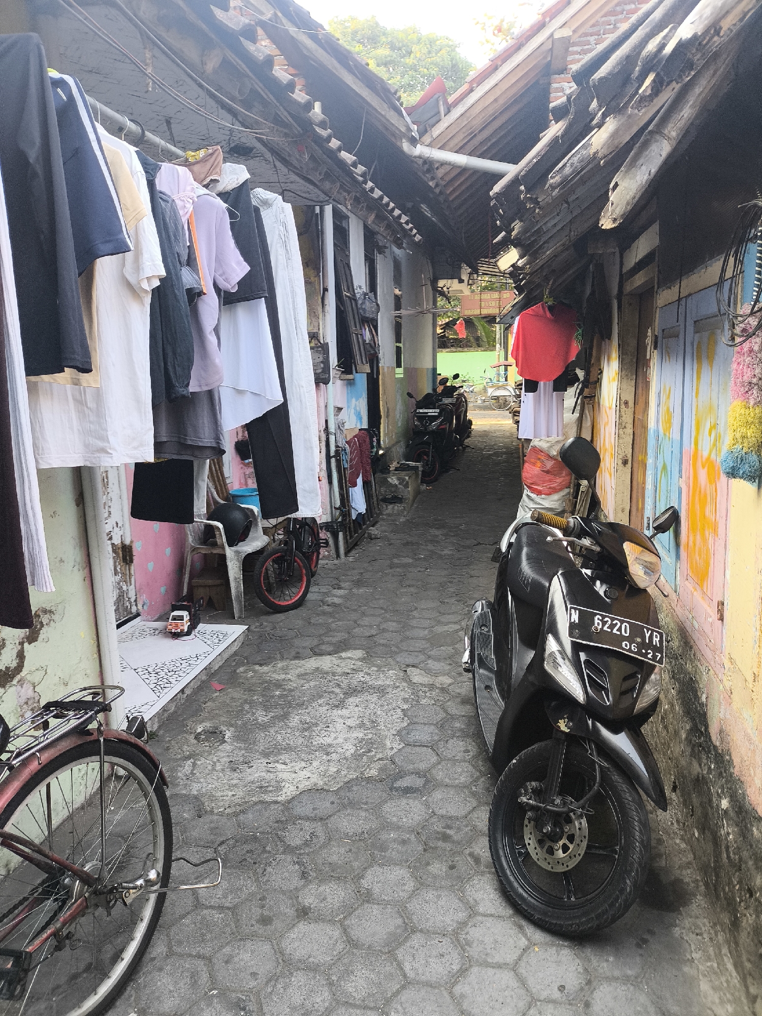 The image depicts a narrow alleyway in the city of Citrodiwangsan, Indonesia. Clothes are hung to dry on a line stretched between buildings. A black scooter is parked on the right side of the alley, its license plate reading "N 6220 YR 06-27". A red bicycle is visible on the left side of the alley. The buildings lining the alley are weathered, with exposed wooden beams supporting tiled roofs. The walls of the buildings are painted in various colors, some showing signs of peeling and wear. The ground is paved with hexagonal stones. The scene suggests a residential area with everyday life activities taking place.