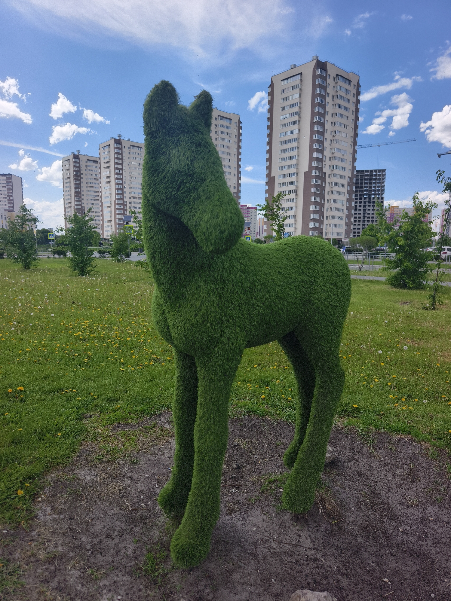 The image depicts a topiary horse in a park-like setting. The horse is covered in green material resembling grass or turf. In the background are several tall apartment buildings under a partly cloudy blue sky. There are some yellow flowers scattered in the grass around the statue. Based on visual details and prior knowledge, it's likely located in Tyumen, Russia.