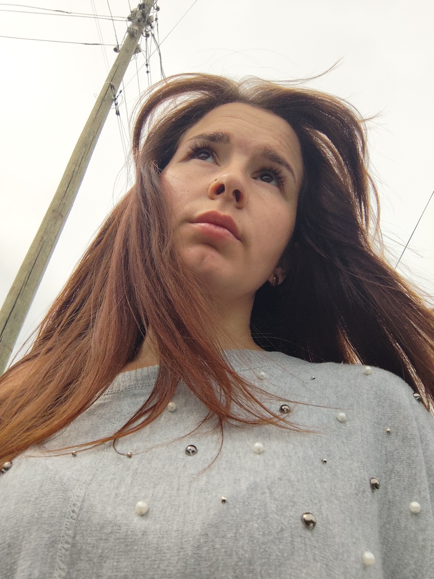 A young woman with long auburn hair stands outdoors, looking up and to the left. Her hair is slightly windblown. She is wearing a light gray sweater adorned with faux pearls and silver studs. In the background, a utility pole with wires is visible against a pale gray or white sky. The photograph appears to have been taken from a low angle. The location is Фоки, Russia.