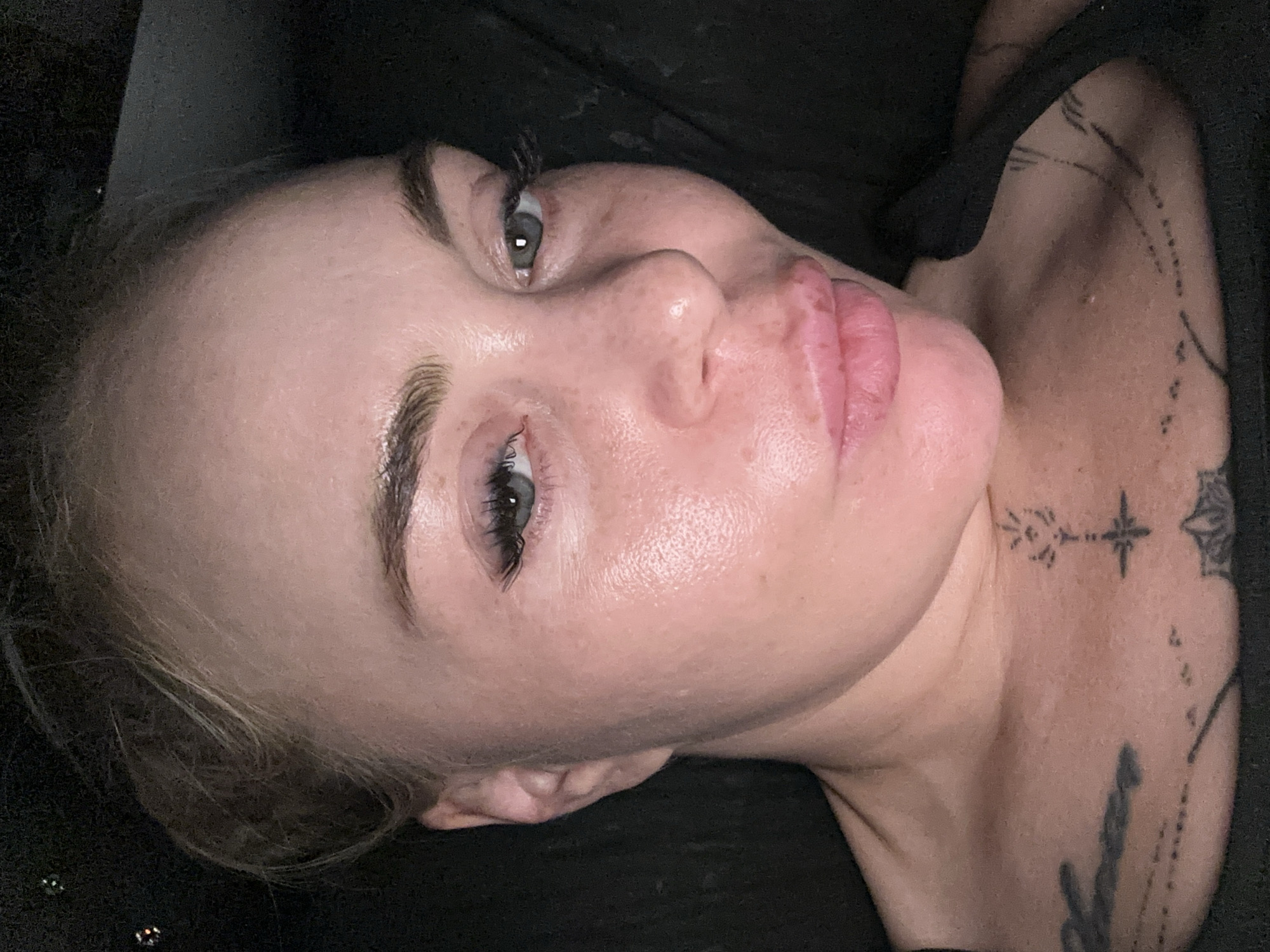 An overhead, close-up shot captures a woman from the chest up. Her blonde hair is pulled back, revealing her face, which is turned slightly to the left. She has a light skin tone with a subtle glow, and her eyes are a light green-blue. Her eyelashes are long and defined, and her eyebrows are filled in. Her lips are full and slightly pursed. There are tattoos visible on her chest and collarbone. She is wearing a black tank top. The background is dark and appears to be a soft surface, possibly a pillow or blanket. The lighting suggests an indoor setting, possibly at night, with soft, diffused light creating highlights on her skin. There are a few small, bright spots in the upper left portion of the frame, which could be reflections or bokeh effects.