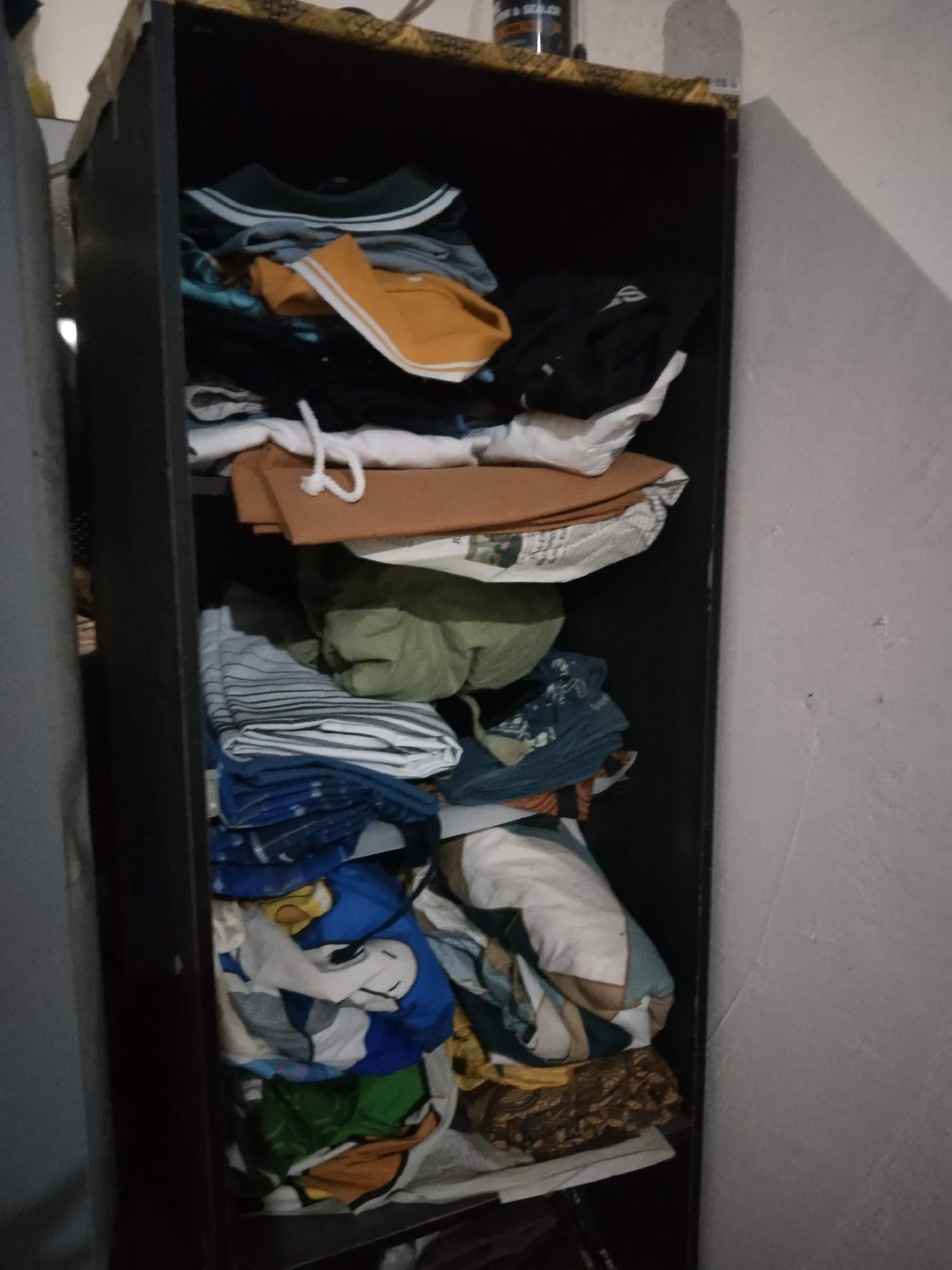In a tall, dark wooden cabinet, a disorganized collection of clothing is stacked on shelves. The items are varied, including t-shirts, pants, and what appear to be towels or blankets. The colors range from muted tones like grey and brown to brighter blues and yellows. Some of the clothing is folded, while other pieces are loosely piled. In the bottom section of the cabinet, a blue t-shirt with a cartoon character design is visible. The cabinet is positioned against a white, textured wall. The image appears to be taken indoors, possibly in a bedroom or storage area in Buntalan, Indonesia.