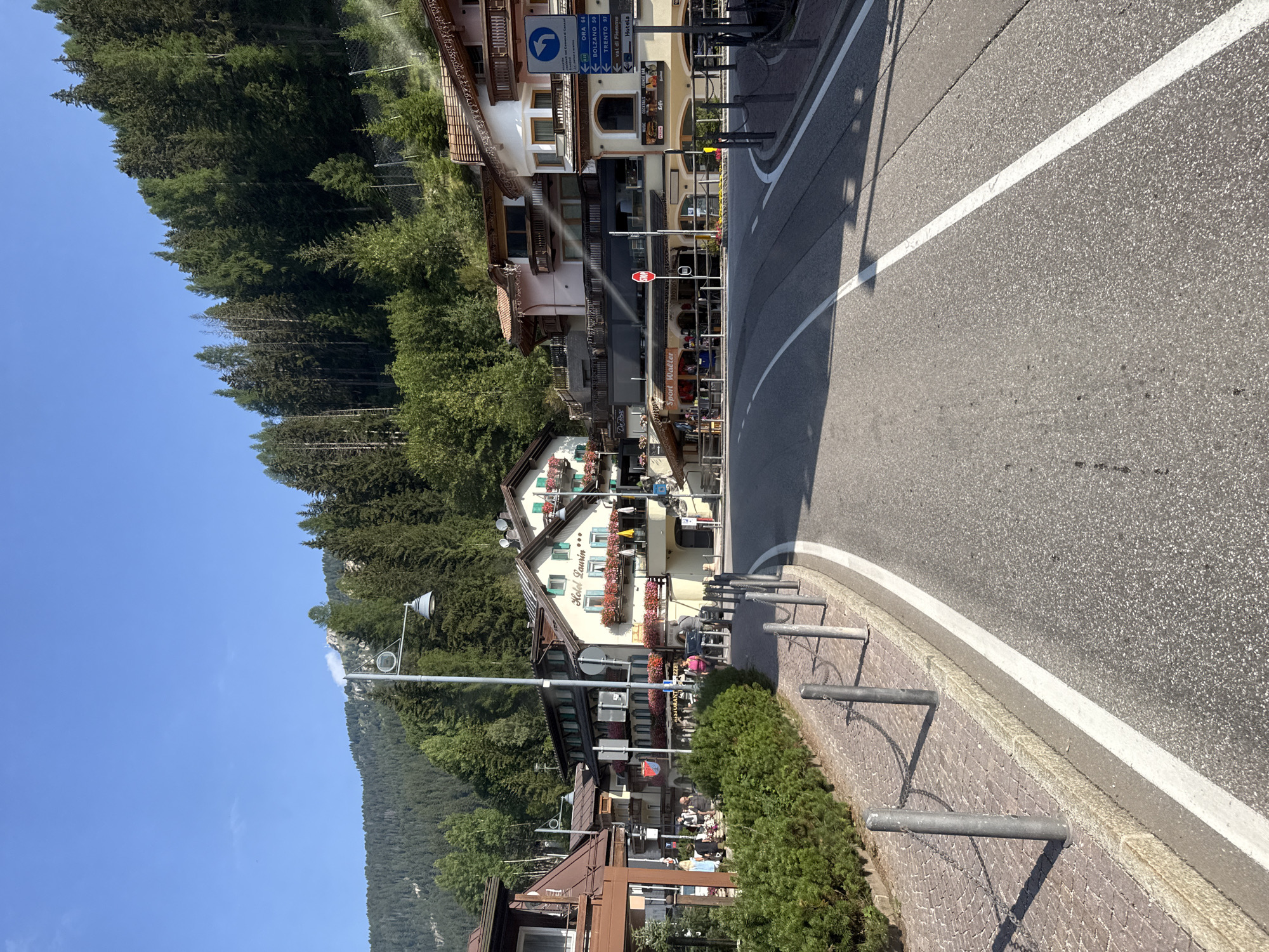 The image depicts a street scene in the mountain town of Canazei, Italy, on a clear day. The road, paved with asphalt and marked with white lines, curves into the distance. On the left, a cobblestone sidewalk is lined with metal bollards and chains, bordered by a dense, green hedge. Several buildings, typical of alpine architecture with wooden balconies and pitched roofs, line the right side of the street. Some of these buildings appear to be hotels and restaurants, with signs indicating "Hotel Laurin," "Ristorante Pizzeria," and "Sport Walter." People can be seen walking on the sidewalk and near the buildings. In the background, a steep, forested mountainside rises, covered in dense evergreen trees. The sky is a clear blue.