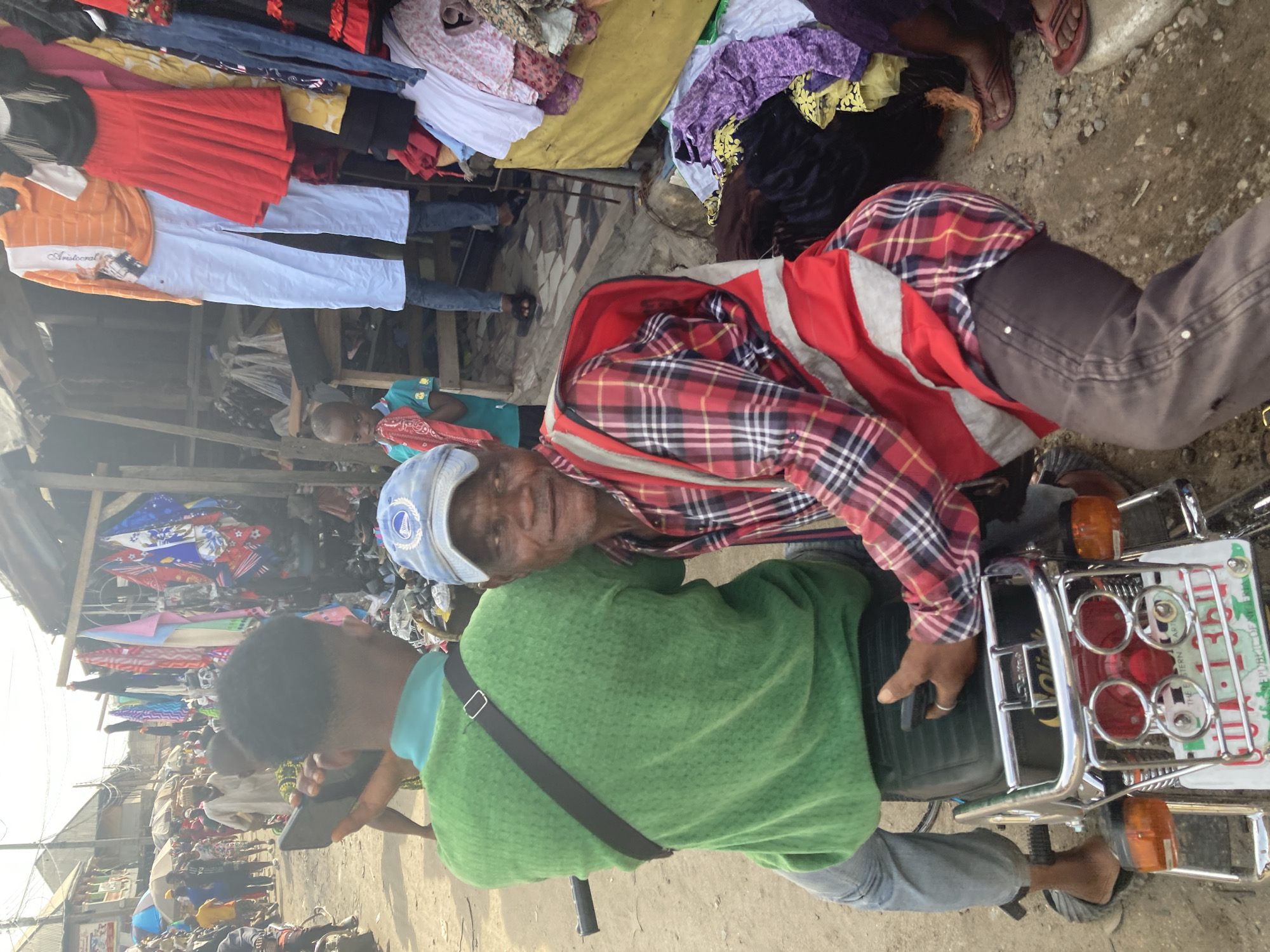 A street scene in Ura Nta Umuarandu, Nigeria, shows a man sitting on a motorcycle holding a cell phone, with another man sitting behind him. The man on the motorcycle is wearing a green sweater and grey pants, while the man behind him is wearing a red and black plaid shirt, grey pants, and a blue hat. A red and white bag is draped across his back. The motorcycle has a metal rack on the back with red circular lights. Clothing is displayed on racks above the men. The background shows other people and stalls, typical of a busy market environment.
