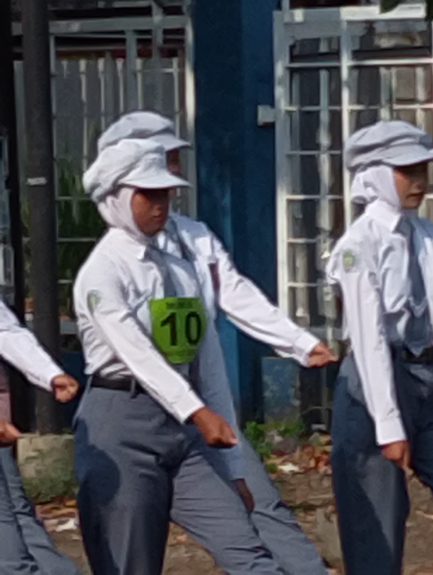 The image shows a person in a white uniform and headscarf, wearing a green bib with the number 10 on it, marching outdoors. They are wearing gray pants and white shoes. In the background, there is a building with windows and a dark blue wall. Another person in a similar uniform is partially visible to the right, also wearing a headscarf and a white shirt. The environment appears to be a sunny day. The location is stated to be in Manaruwi, Indonesia.