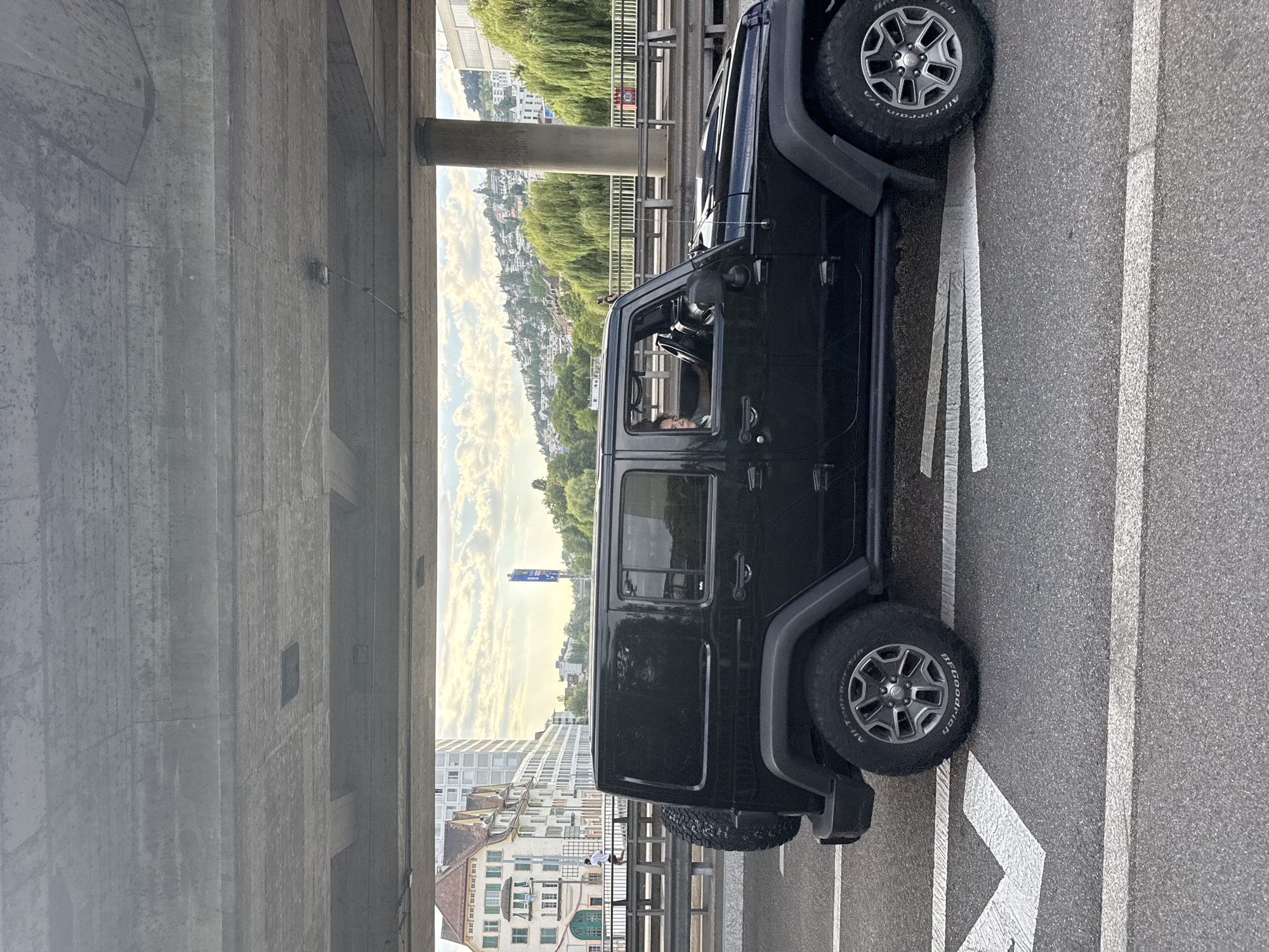 A black Jeep Wrangler is driving on a road under a concrete overpass in Zurich, Switzerland. The Jeep is in the right lane, with white directional arrows painted on the asphalt road. In the background, there are buildings, trees, and a cloudy sky. A person is visible in the driver's seat of the Jeep. To the left of the road, a bridge with railings and a sidewalk is visible, with a person running on the sidewalk. The concrete overpass has a rough texture and appears to be an elevated roadway or bridge. The sunlight suggests it is late afternoon or early evening.