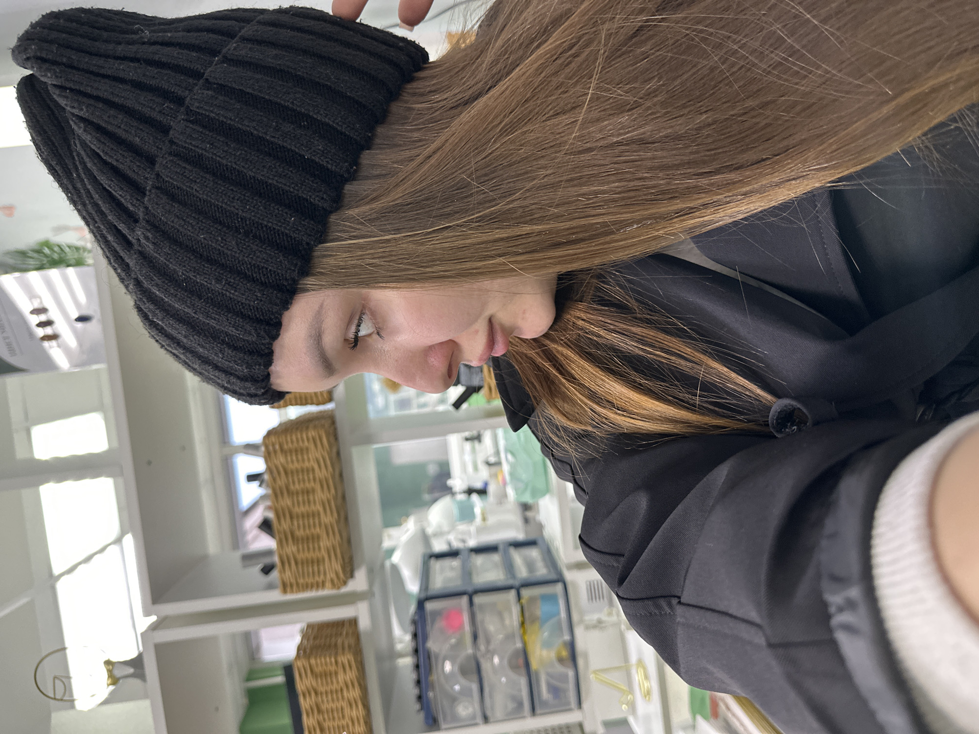 The image, originally rotated 90 degrees counter-clockwise, depicts a young person in an indoor setting, viewed from their right side. The person, likely female, has long, straight, light brown hair cascading over their shoulders and back. They wear a black ribbed knit beanie, with a finger subtly touching the top of it, and a black jacket. Their gaze is directed downwards, with visible eyelashes and the contour of their cheek and lips suggesting a focused or contemplative expression.
The background reveals a brightly lit, organized room, characteristic of a classroom, workshop, or creative studio. On the left, white shelving units hold various items, including a green plant in a white pot. Towards the right and mid-ground, additional white shelves display multiple woven wicker baskets. Prominently featured in the mid-ground is a transparent, multi-compartment storage unit, likely containing small supplies or components. Bright natural light enters from a large window in the background, indicating it is daytime. The overall environment suggests a functional space. No discernible text is visible in the image. The location context is Chaikovsky, Russia.