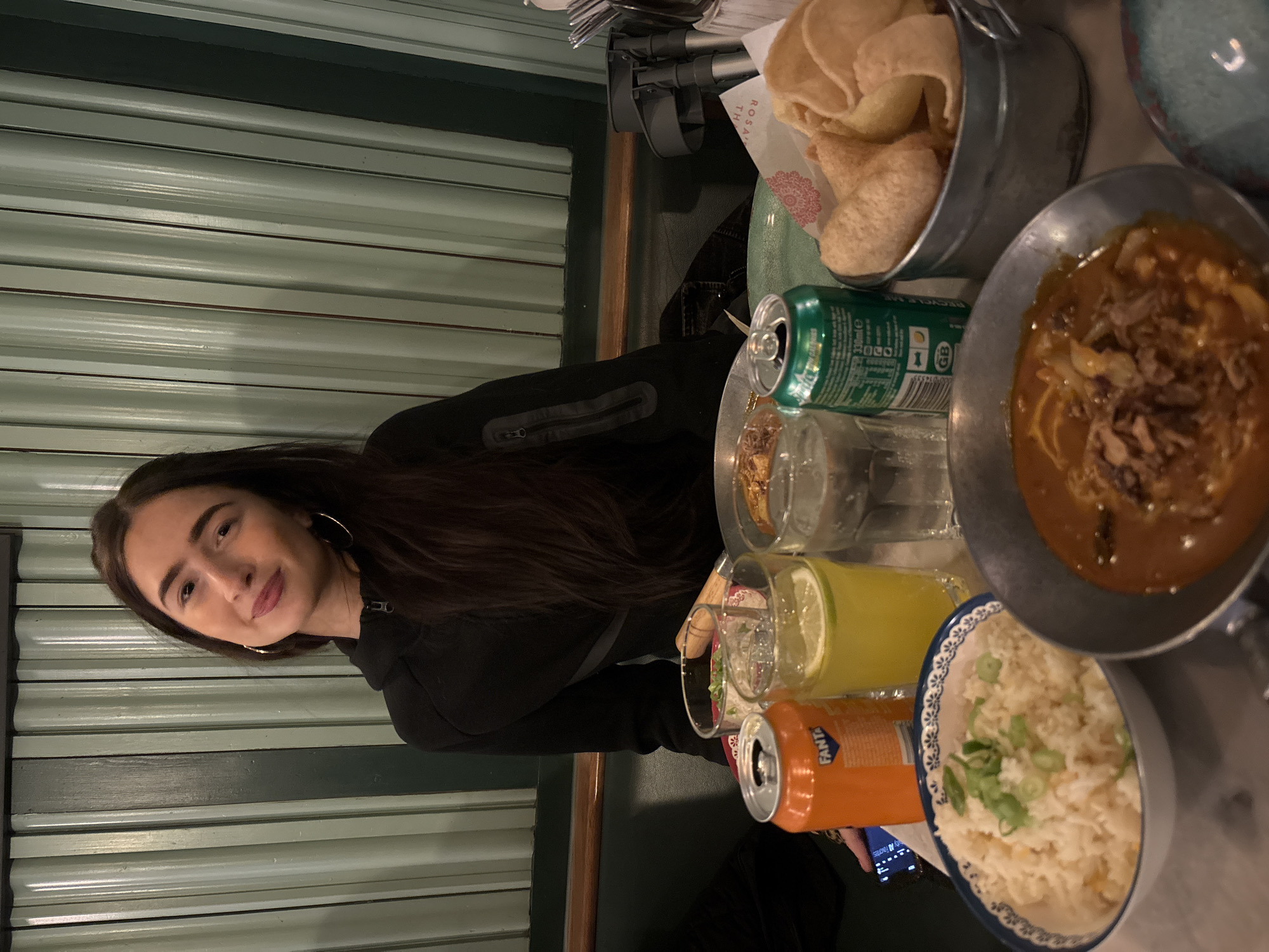A young woman with long brown hair and hoop earrings sits at a table. She is wearing a black jacket. The table is filled with food and drinks, including crackers in a metal container, a dish of curry, a bowl of rice, a glass of yellow liquid with a lime slice, a can of Fanta, and a can of what appears to be soda. The background is a green wall with horizontal lines. The location is likely a restaurant in London, United Kingdom, given the style of the dishes and drinks.