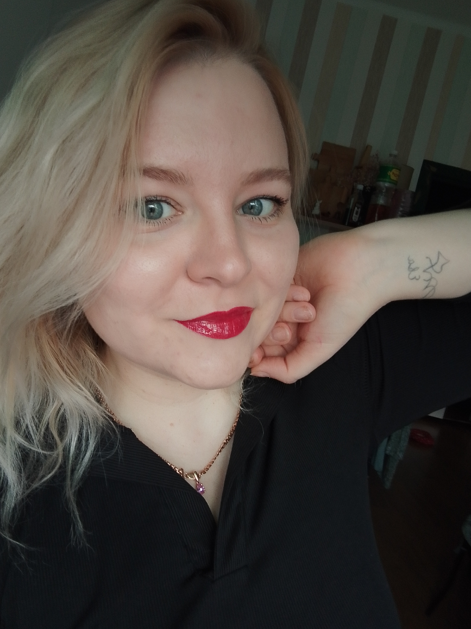 A blonde female subject with blue-green eyes and red lipstick is depicted smiling and looking directly at the viewer. She wears a black ribbed top and a gold-colored necklace with a small pink gemstone pendant. Her left hand is positioned against her lower face/neck. A faded, dark-ink tattoo with abstract or stylized script is visible on the inner aspect of her left forearm/wrist. The background is an out-of-focus indoor environment featuring a vertically striped wall. Visible on a surface behind the subject are miscellaneous objects, including bottles, a wooden box, and a dark electronic device. No visual information within the image confirms the specified location as Izhevsk, Russia.