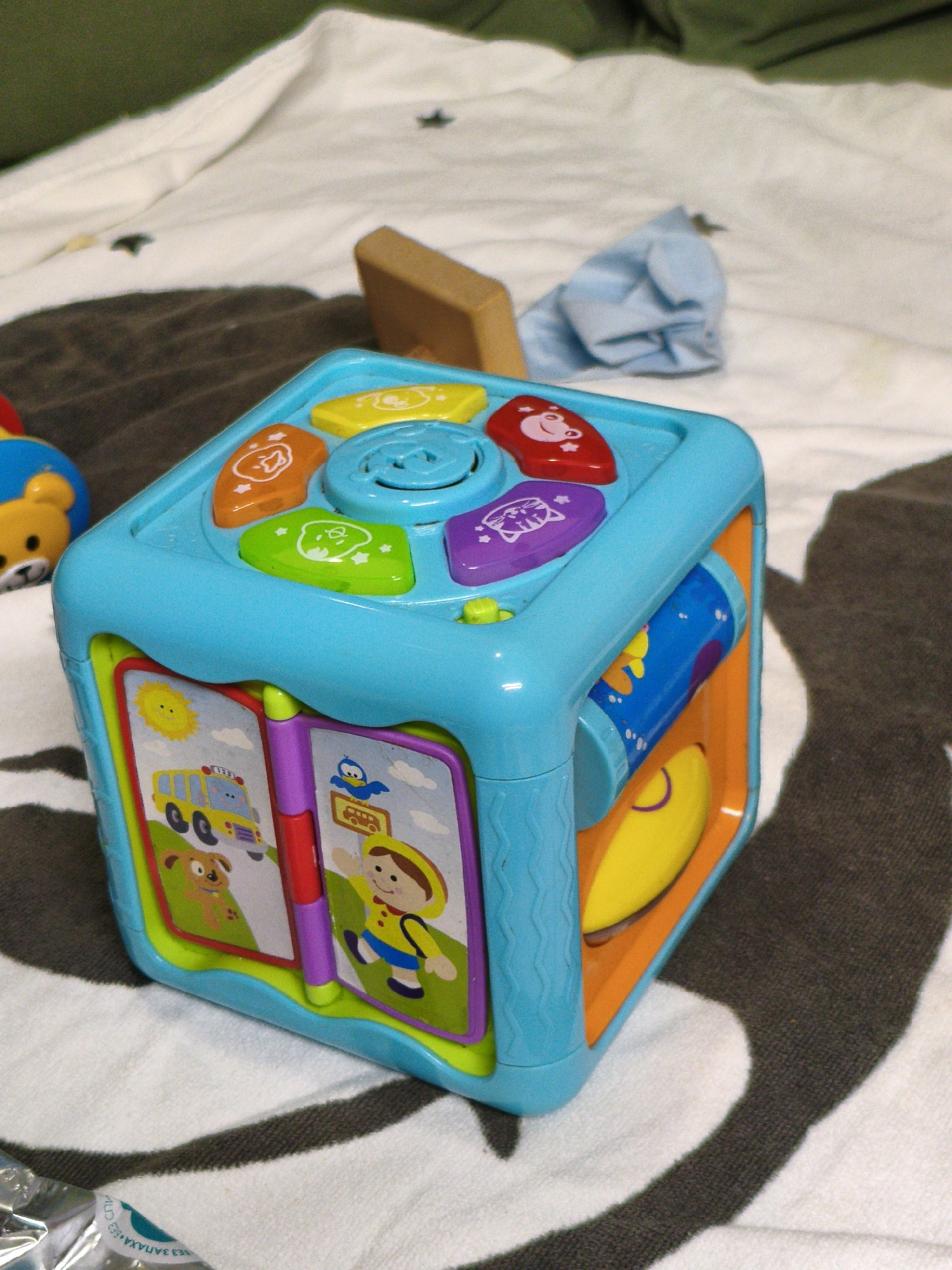 A bright blue toy cube, featuring various interactive elements and colorful illustrations, rests on a white blanket with black stripes. The toy has several buttons on its top surface, including orange, green, and purple ones adorned with cartoonish animal faces. One side of the cube depicts an open book with images of a yellow school bus, a dog, a smiling sun, and a cartoon child with a blue backpack and yellow shirt. Another side shows a spinning cylinder with blue and yellow patterns. The background consists of a dark surface and a green fabric, possibly a bedspread. The context of Lyubertsy, Russia, is not evident in the provided image.