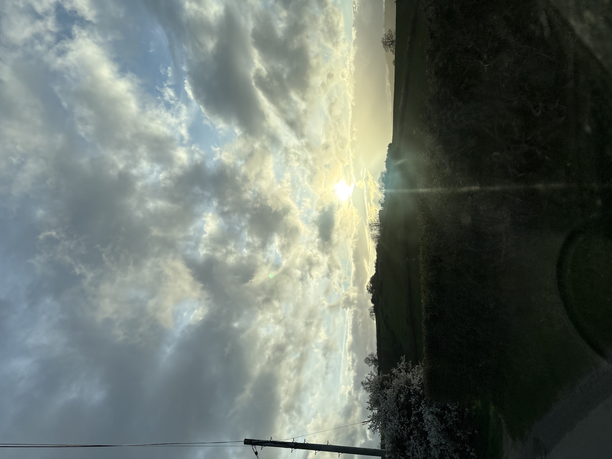 The image depicts a cloudy sky with the sun shining through the clouds, along with a view of the surrounding landscape. The scene includes a field, hedgerows, trees, and utility poles. The location is likely in a rural or semi-rural area near Shrewsbury, United Kingdom. There are no people visible in the image. The sun is the primary object of focus, with its rays shining through the clouds. There are no interactions or actions visible other than the natural environment.