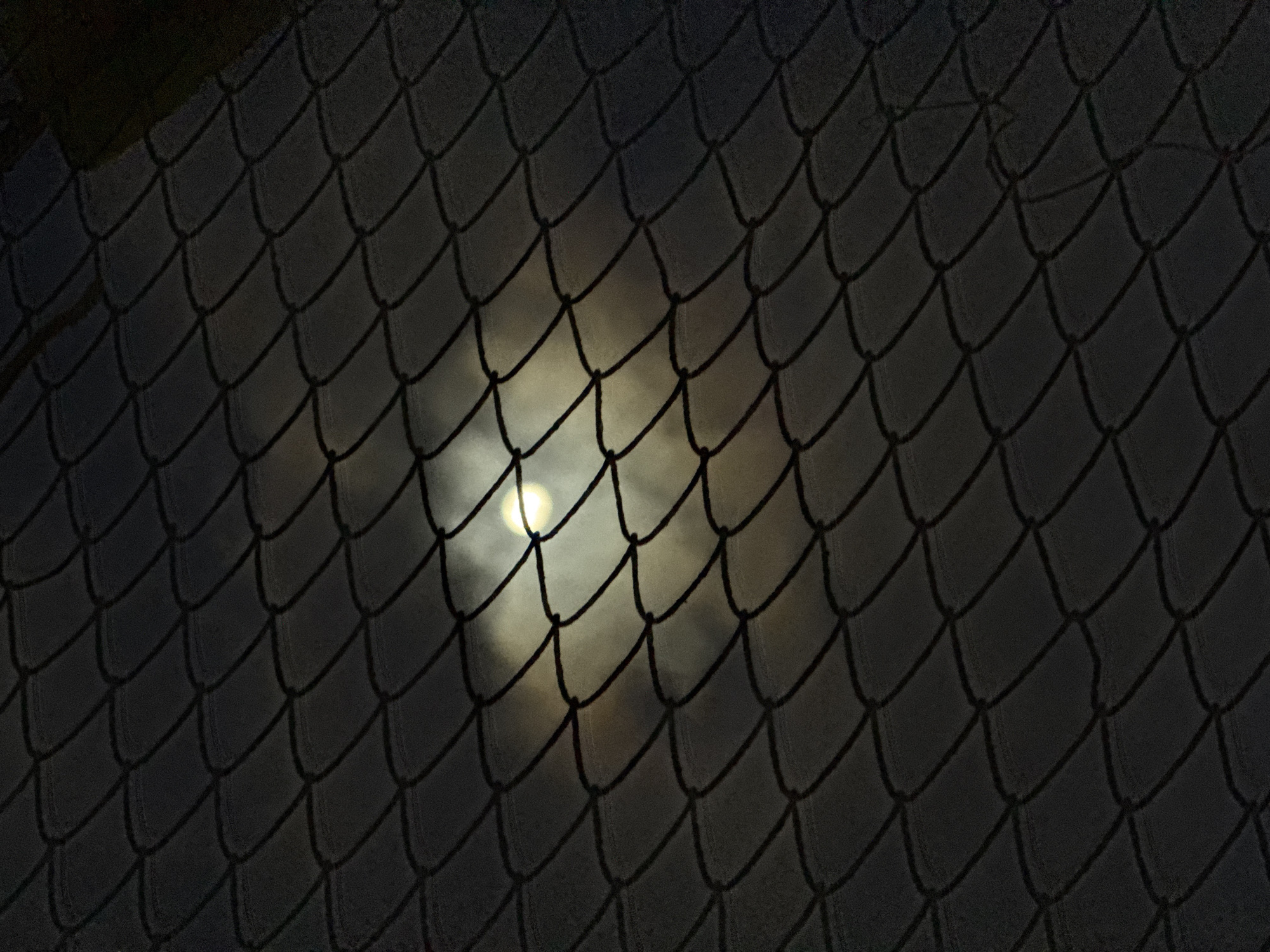 A nighttime photograph, taken in Denpasar, Indonesia, captures the moon peeking through clouds, viewed through a diamond-patterned chain-link fence. The fence, dark and out of focus, dominates the foreground, its metallic weave creating a grid-like effect. Behind it, the moon appears as a bright, luminous orb, partially obscured by wispy, grey clouds. The overall mood is one of mystery and urban observation, with the fence acting as a barrier between the viewer and the celestial display. The scene is devoid of human presence.