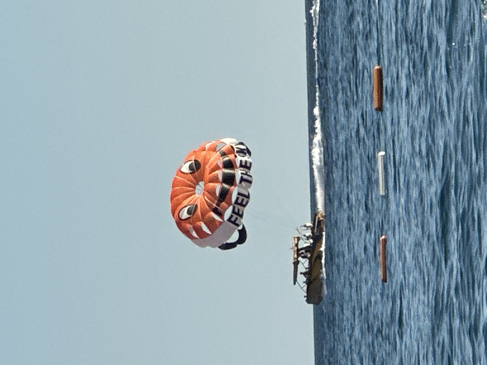A parasail with a cartoon face and the words "FEEL THE" visible is airborne above a body of water. Below the parasail, a motorboat with several people on board is moving through the water, creating a wake. The water is blue and choppy, with some buoys floating on the surface. The sky is clear and light blue. The scene takes place in the city of Boreti, Montenegro.