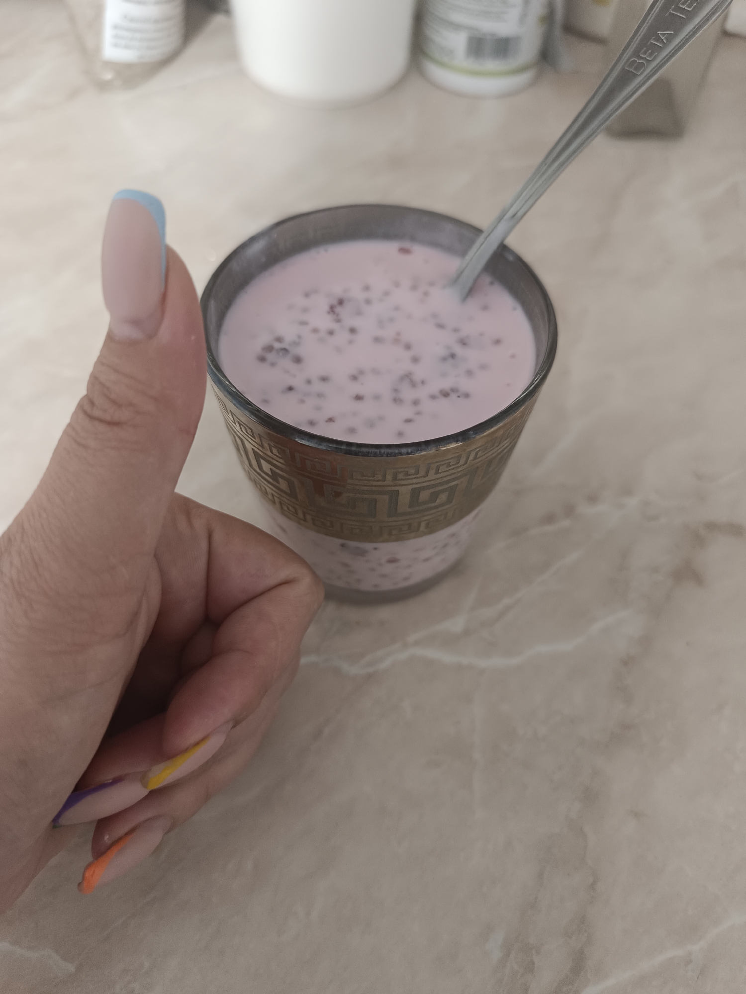 A close-up, slightly elevated shot shows a person's left hand giving a thumbs-up next to a glass of pink liquid with dark specks, possibly chia seeds or berries. The glass has a gold decorative band near the top. A spoon rests in the glass, with the engraving "BETA TE" visible on its handle. The background consists of a beige, marbled countertop. The person's fingernails are manicured with different colors: light pink with blue tips on the thumb, and orange, yellow, and purple designs on the other visible nails. The image's lighting suggests an indoor setting.
