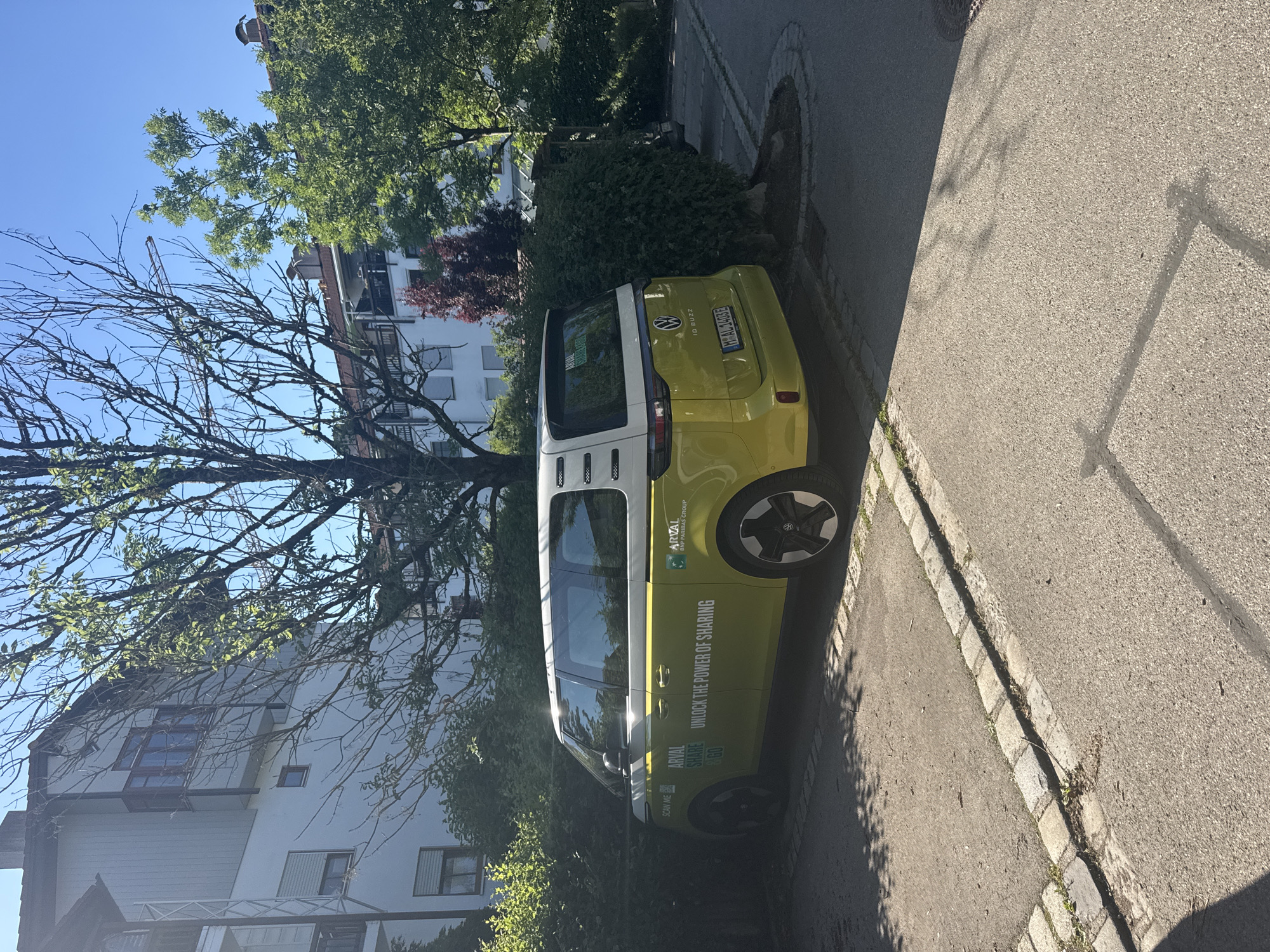 The image depicts a bright, sunny day in a residential area, likely in Höhenkirchen-Siegertsbrunn, Germany. In the foreground, a bright yellow Volkswagen ID. Buzz, branded with "ARVAL SHARE UNLOCK THE POWER OF SHARING," is parked on a paved surface bordered by cobblestones. The car is positioned adjacent to a dense green hedge. Behind the hedge and car, white apartment buildings with balconies and windows are visible. Trees with green foliage and bare branches are scattered throughout the scene, with a small red-leafed tree also present. A construction crane is visible in the distance, partially obscured by trees and buildings, suggesting ongoing development in the area. The ground is a mix of asphalt and gravel. The license plate of the car is visible and reads "M AL 3405 E".