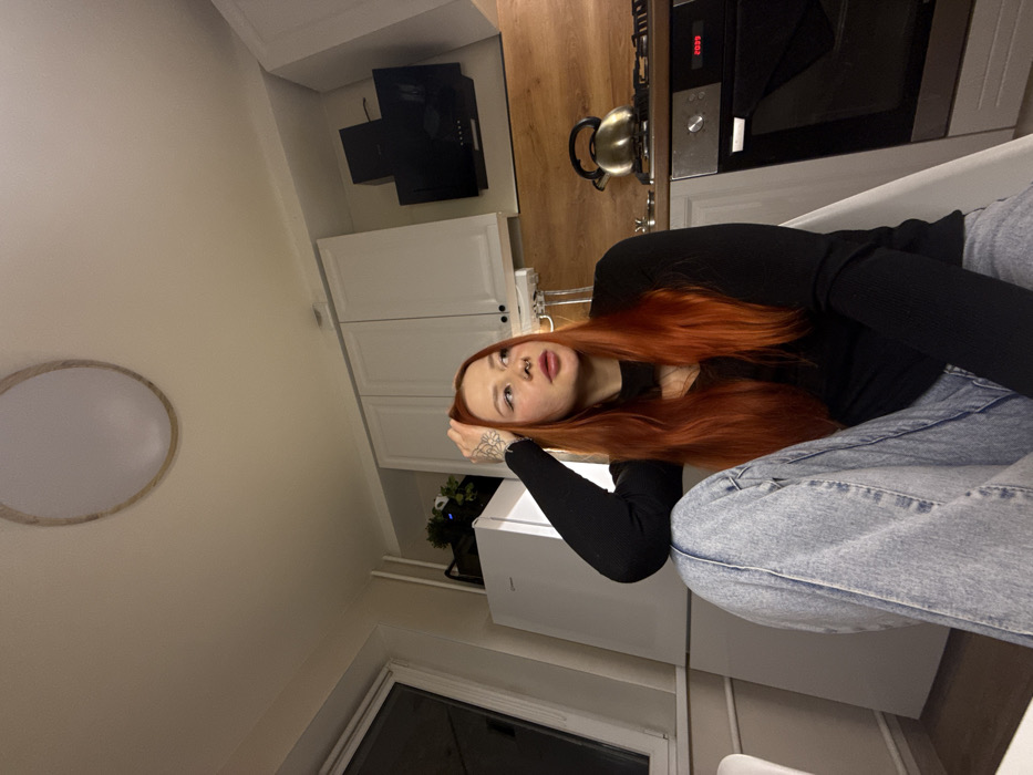 The image captures a young woman reclining in what appears to be a modern kitchen setting. The image is oriented sideways, with the woman lying on her back, looking up towards the viewer, and the kitchen elements arranged vertically behind her.
The woman has long, reddish-orange hair that cascades around her shoulders. She is wearing a black, long-sleeved top and light blue denim jeans. Her left arm is bent, and her hand is resting near her head, revealing a tattoo on her wrist or forearm, which appears to be a floral design. She has a nose piercing (a stud in one nostril) and her lips are slightly parted. Her gaze is directed upwards, towards the camera.
The background shows a kitchen area. To the woman's right (when viewed correctly), there is a section with white upper and lower cabinets. Above the countertop, a black range hood is visible, and below it, an oven or stove unit is integrated. The oven has a digital display showing "2039", likely indicating the time. A metallic kettle with a dark handle is resting on the wooden countertop surface next to the oven.
Further to the left of the woman (when viewed correctly), there is a large white appliance, possibly a refrigerator or a dishwasher. Behind it, a small potted plant with dark green foliage can be seen. The wall is a light, neutral color. In the upper left corner, a large, round, flush-mount ceiling light fixture with a white diffuser and a light-colored rim is visible. A portion of a window frame is also visible in the lower left corner of the image.
The overall impression is an informal, personal snapshot taken indoors, likely in an apartment or house kitchen. The lighting suggests it might be evening or nighttime, consistent with the time displayed on the oven.