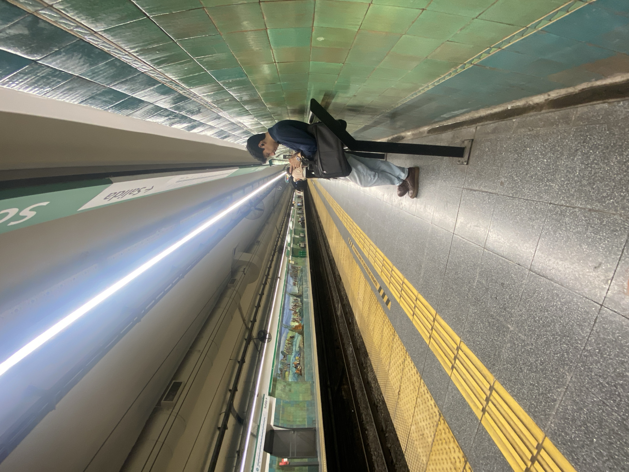 The image shows a man standing on a subway platform in Buenos Aires, Argentina. He is leaning against a black metal barrier. He is wearing a dark blue shirt, light-colored pants, and brown shoes, and carrying a dark-colored bag. The platform has green tiled walls and a grey tiled floor with yellow tactile paving. The train tracks are visible to the left of the platform, as well as a train with murals along its side. A sign indicating "Salida" (Exit) is also visible. The lighting in the station is bright, with fluorescent lights running along the ceiling.